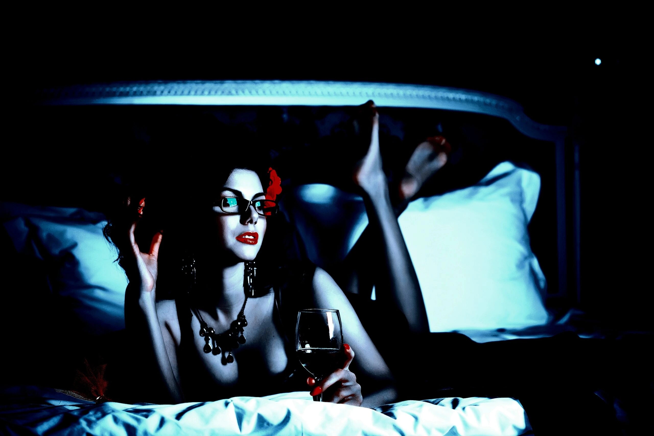 A glamorous woman in glasses and dark attire lounges on a bed, holding a glass of wine, combining elegance with a sultry, enigmatic look.