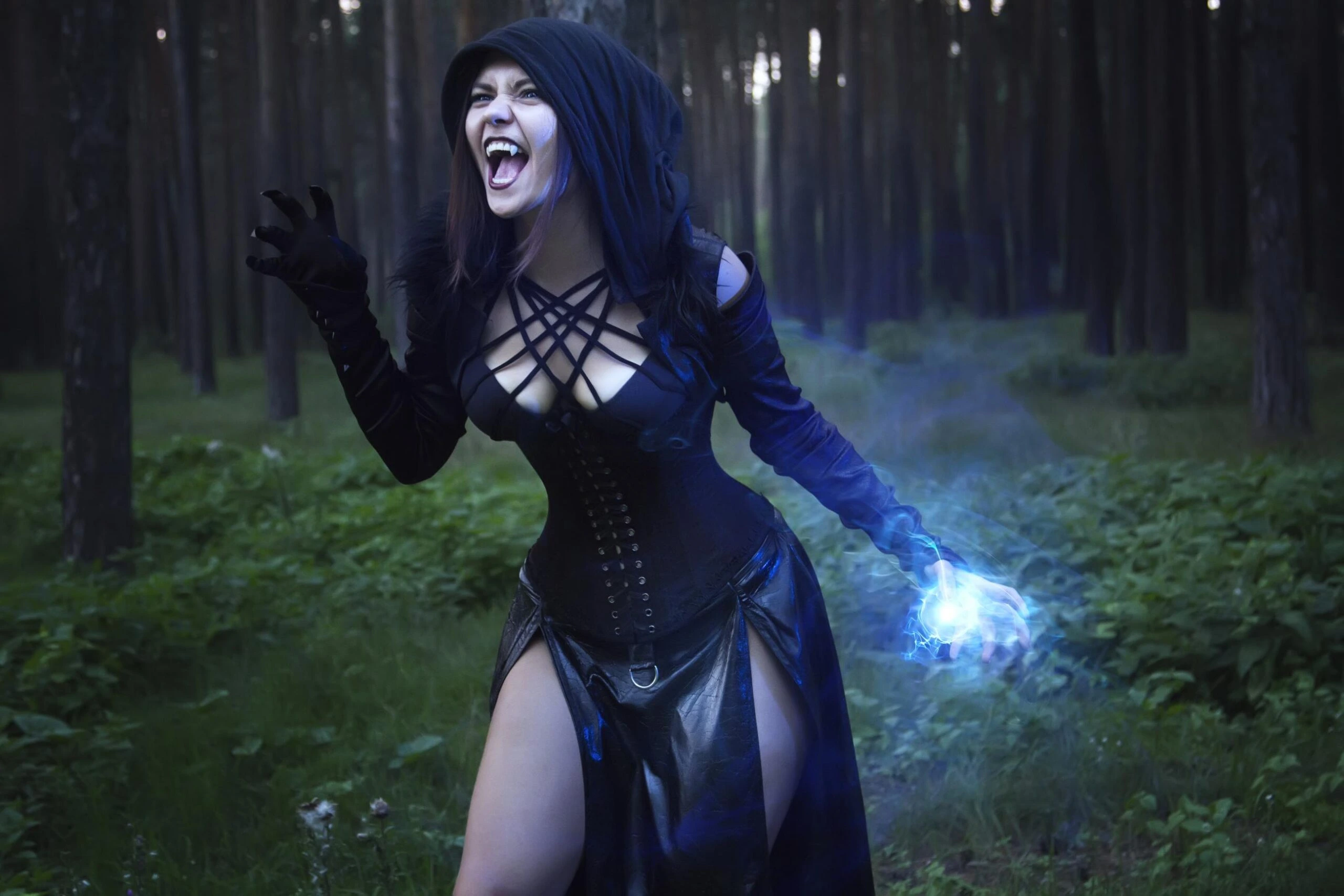A dark sorceress cosplayer in a revealing black outfit with a hood, casting a blue magical spell in a dense forest at twilight.
