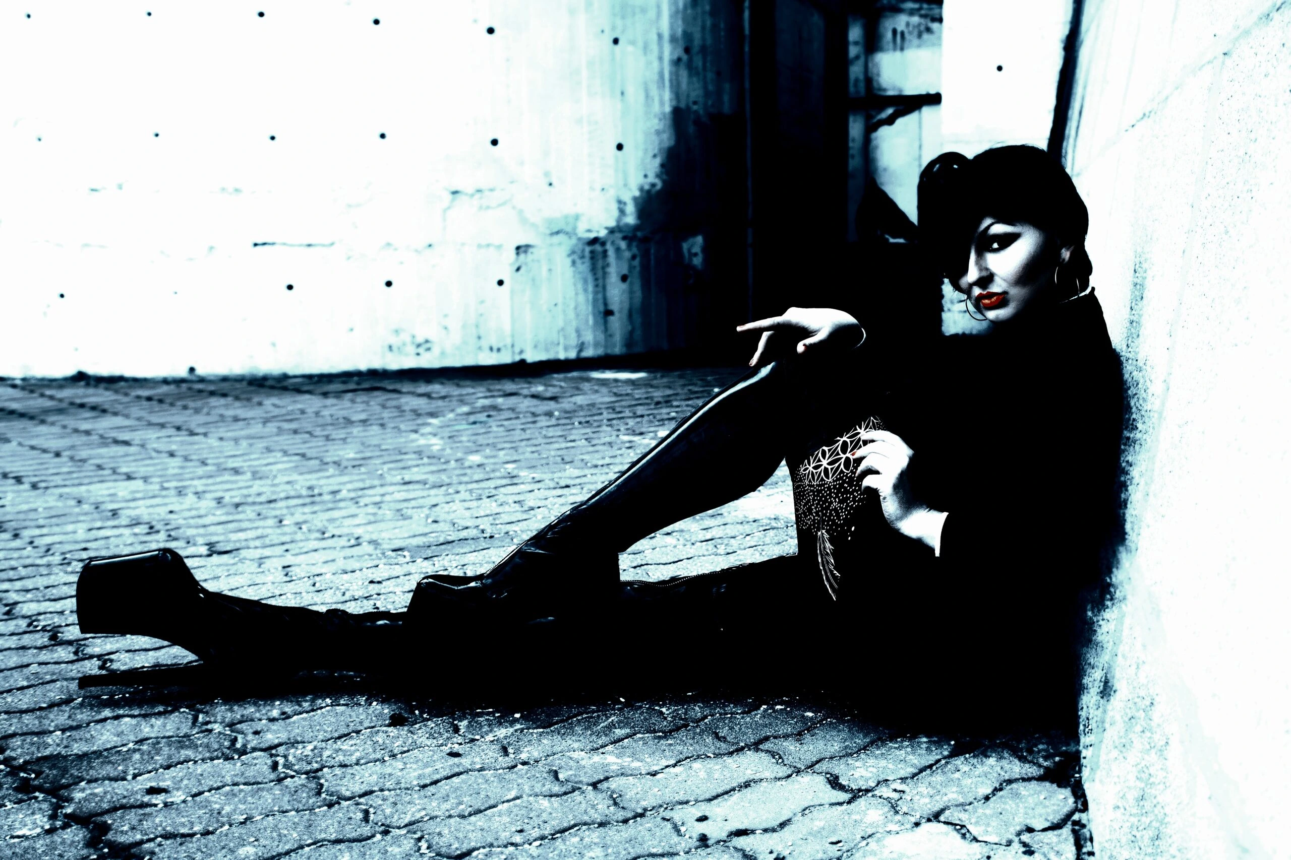 A woman dressed in dark, gothic attire sits against a concrete wall, exuding a moody and edgy vibe with bold red lipstick and high platform boots.