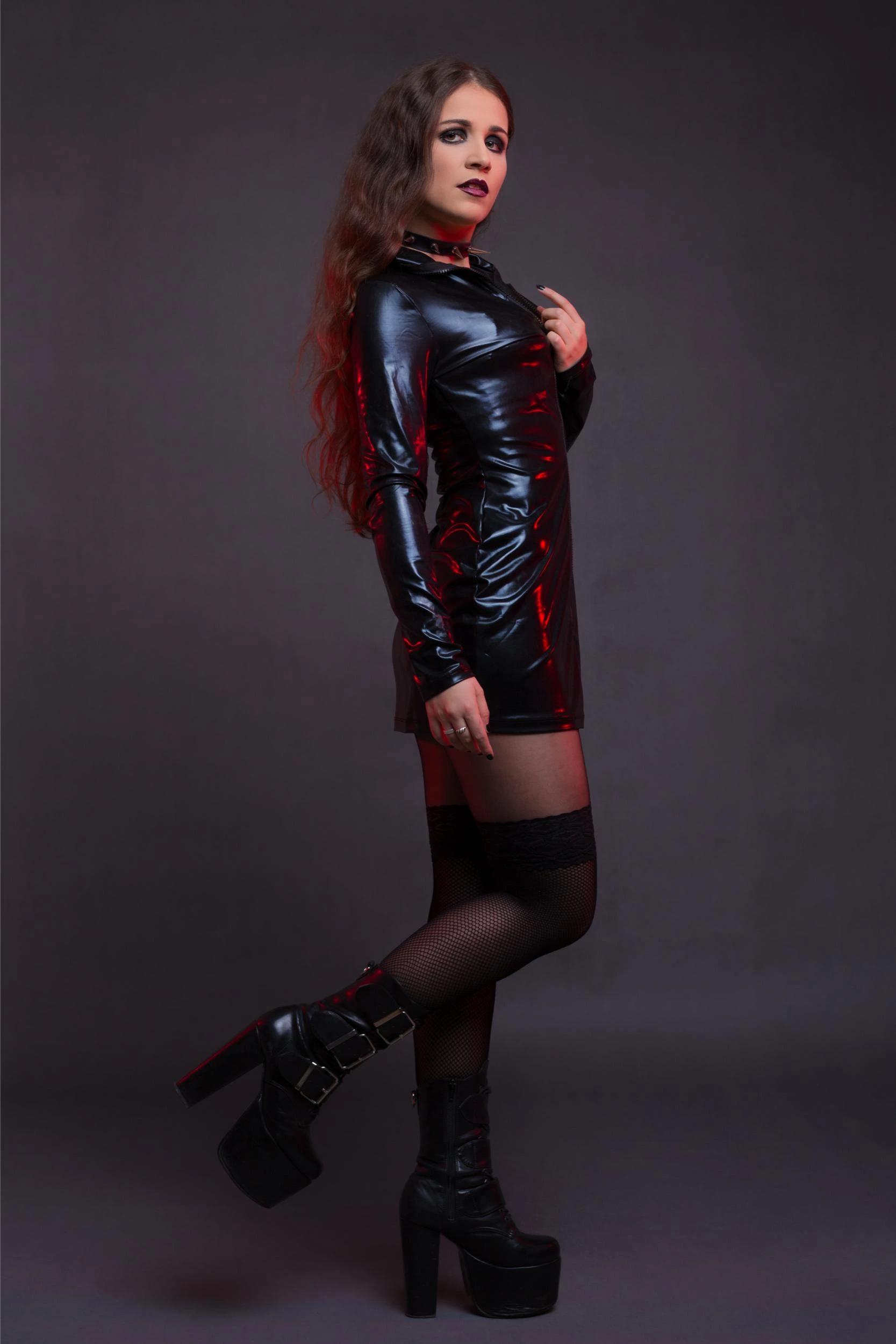 A woman dressed in a sleek, shiny black latex dress with a high neckline, dark makeup, and platform boots, posing confidently against a dark background.