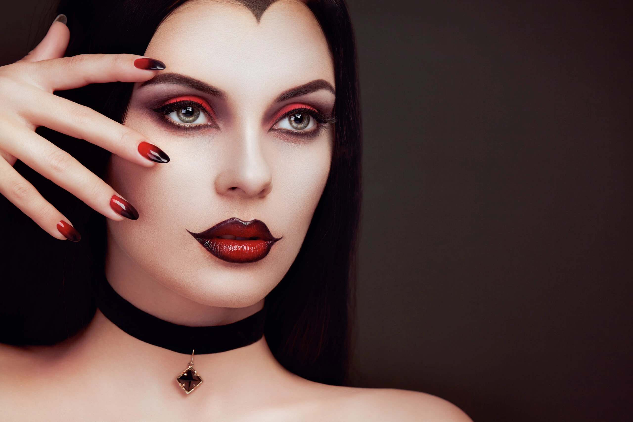 Close-up of a woman with a gothic look, featuring sharp red nails and dark makeup, wearing a black choker with a gemstone pendant.