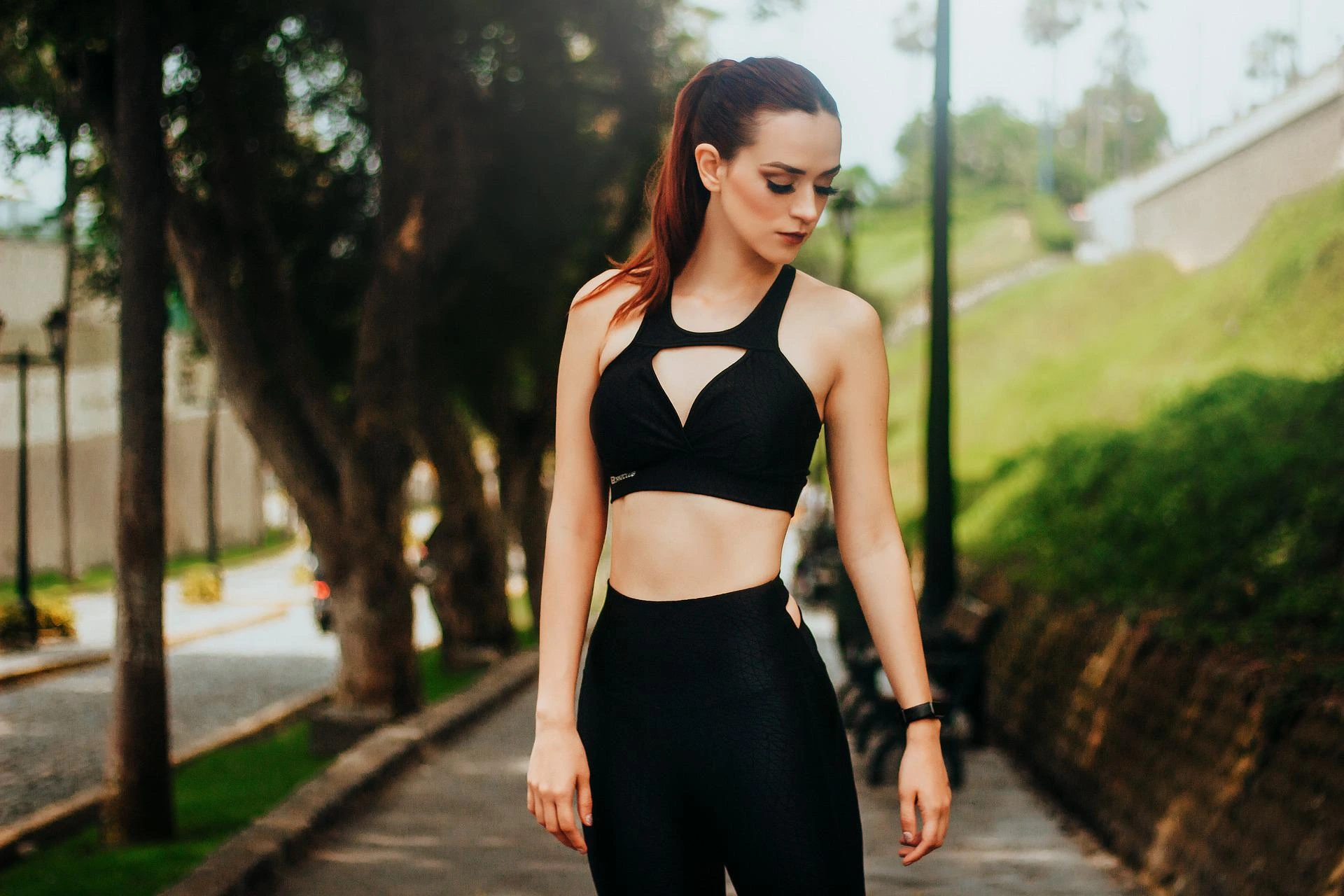 A woman in black fitness wear standing outdoors, with trees and a park path in the background, exuding confidence.