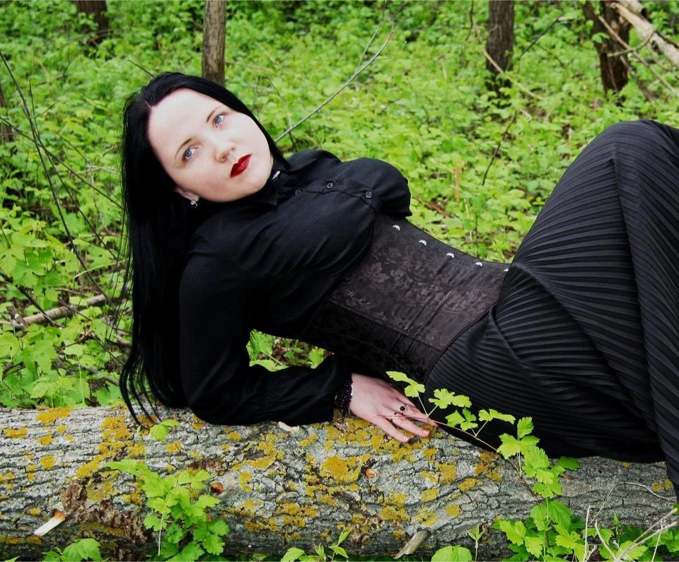 Woman in black underbust corset and dark flowing dress reclining on a mossy tree trunk in a lush green forest.