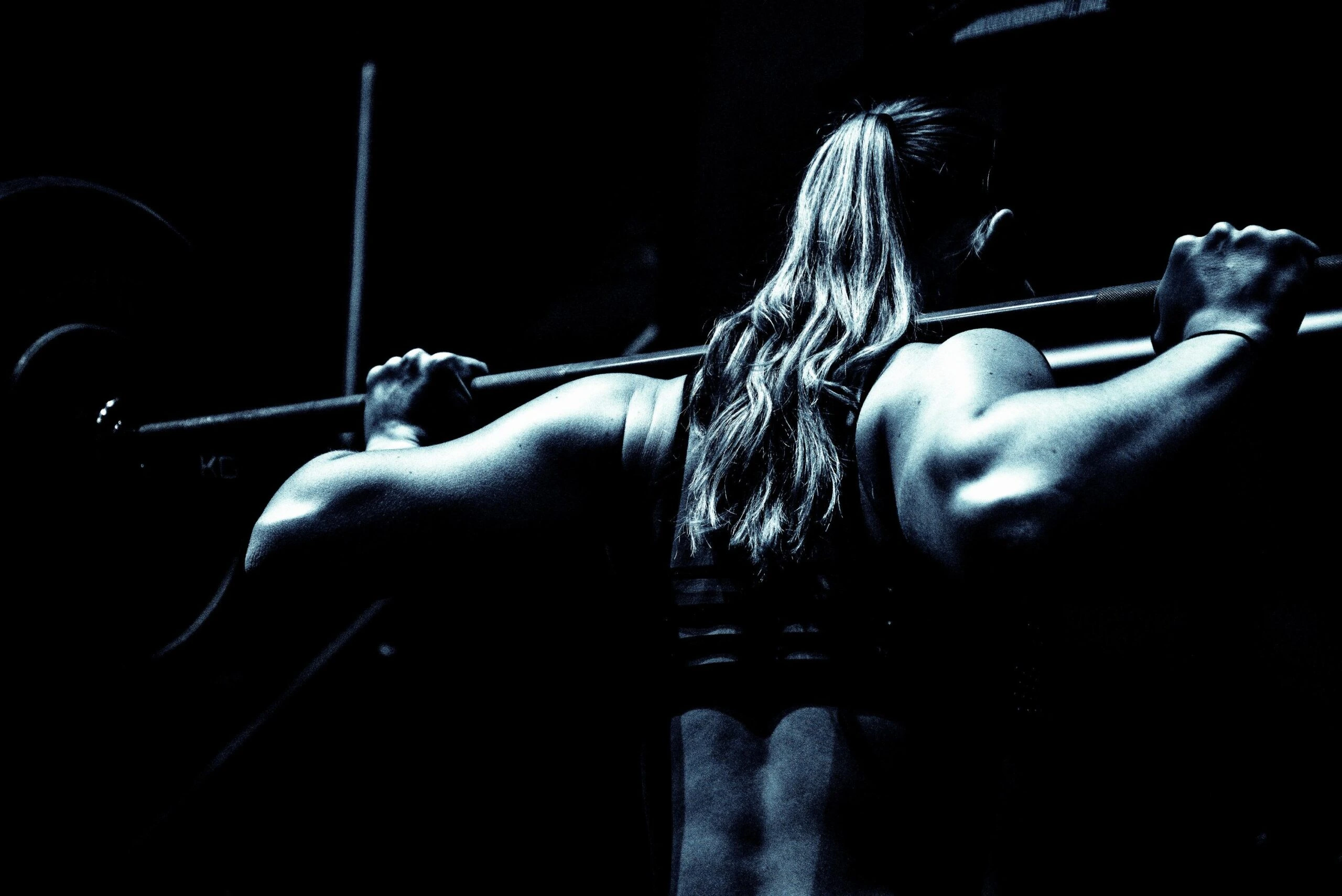 Back view of a woman lifting a heavy barbell in a gym, her muscular arms emphasized by dramatic lighting.