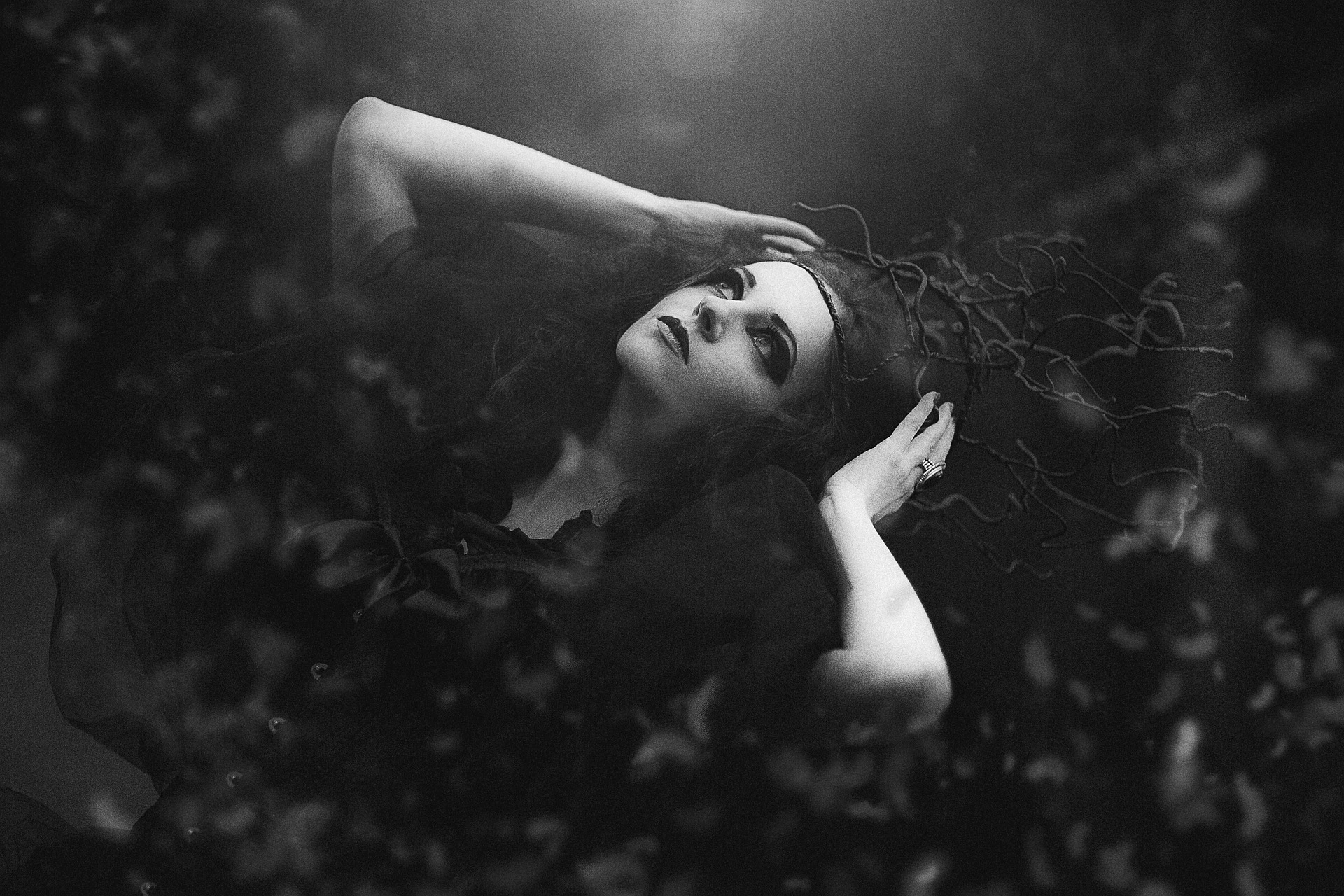 Ethereal black and white portrait of a gothic woman with dark makeup, lying with her hands in her hair adorned with twisting branches.