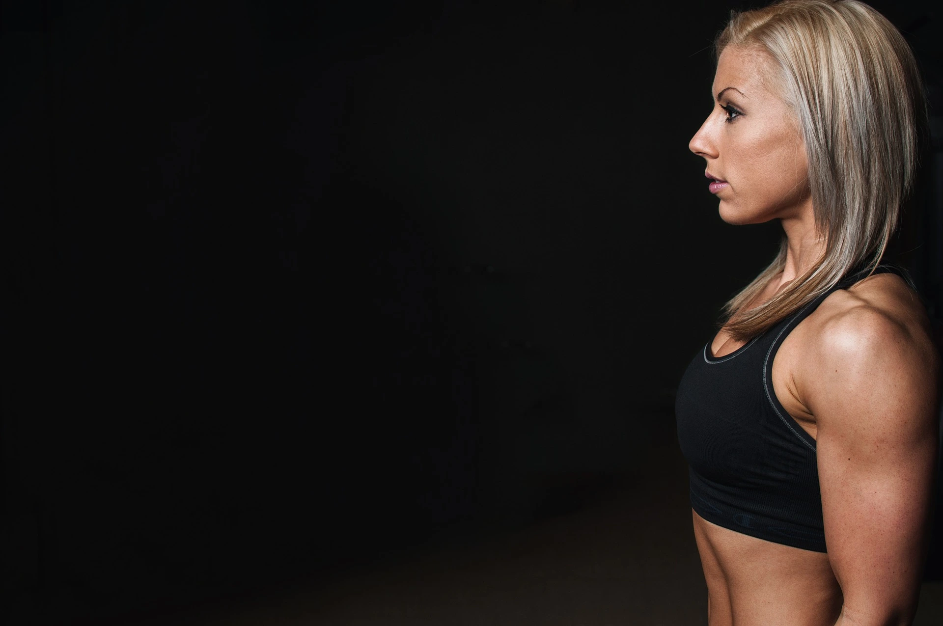 A fit woman in a black sports bra standing in profile against a black background, highlighting her toned arms and physique.