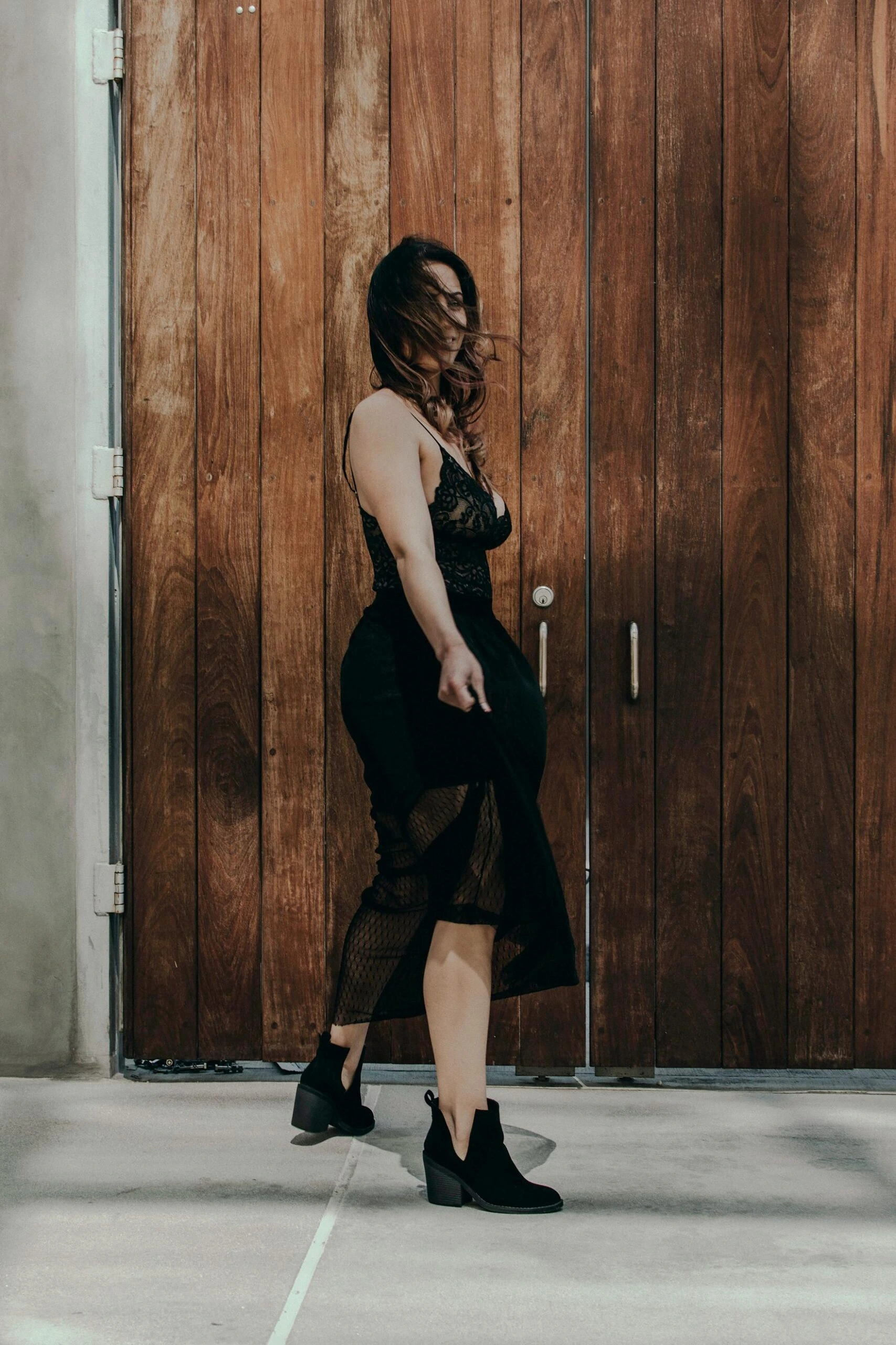 Woman walking past a wooden door wearing a black lace dress with mesh panels, her hair blowing in the wind.