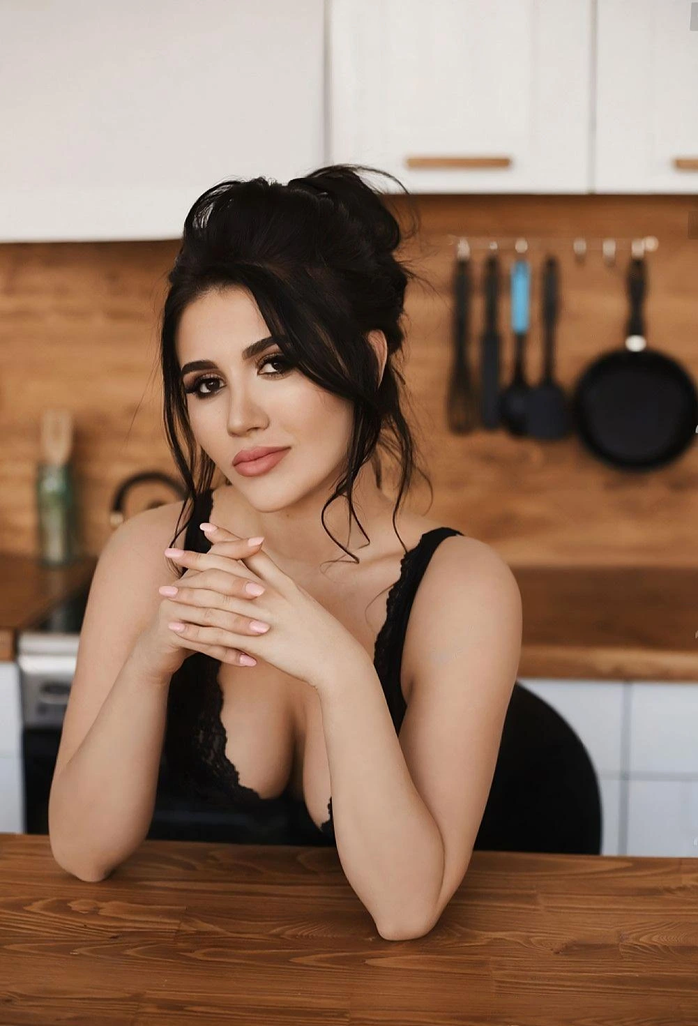 Woman with dark hair in a kitchen, hands clasped together, giving a calm expression.