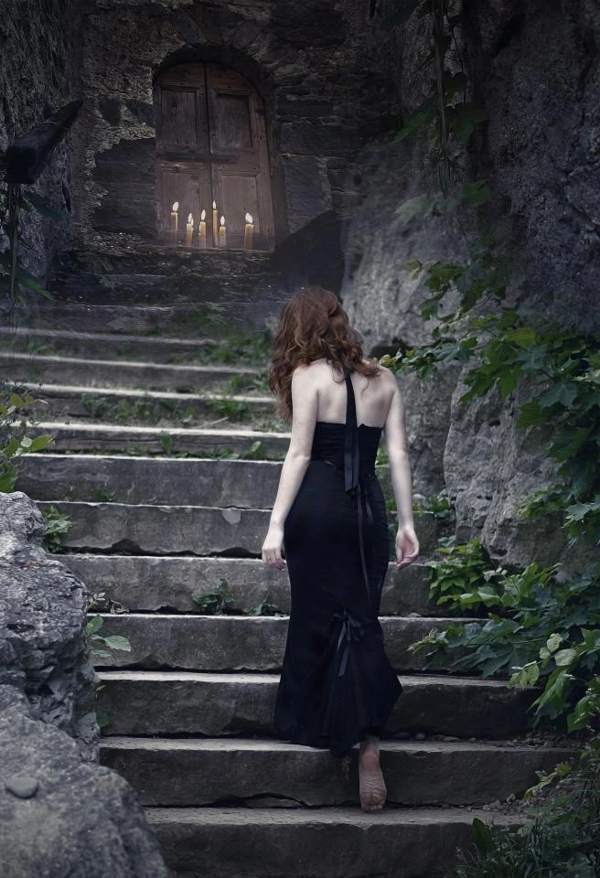 Woman ascending stone stairs in a flowing black dress, heading toward a mysterious doorway lit by candles.