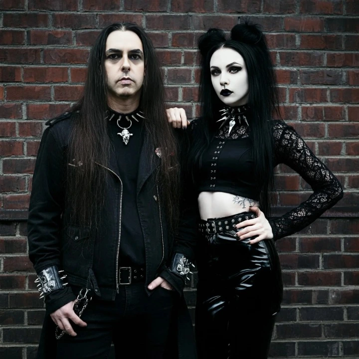 Black metalhead man and gothic woman in dark clothing, standing against a brick wall, with an intense, rebellious expression.