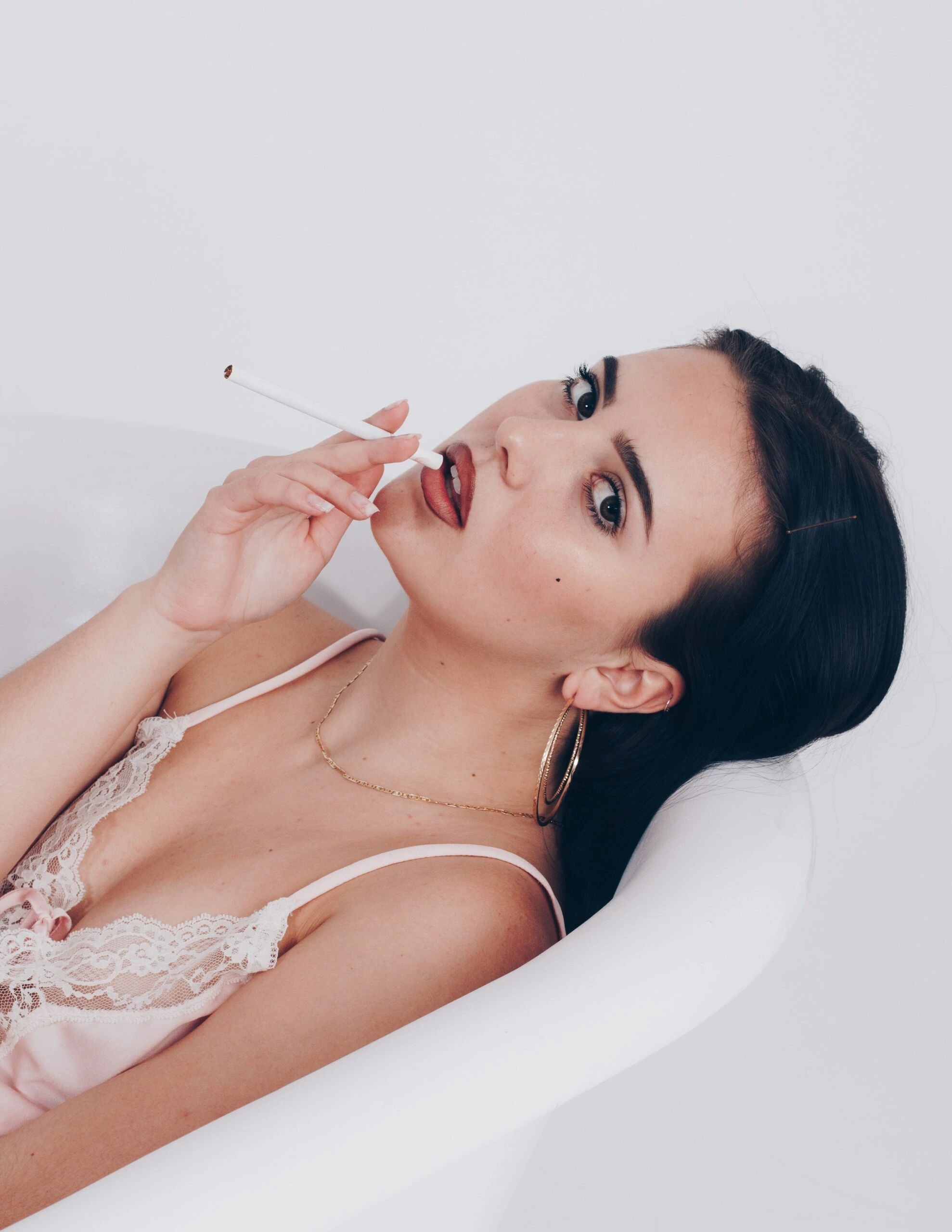 Woman in a pink lace nightgown, lounging in a white bathtub and holding a cigarette with a sultry expression.