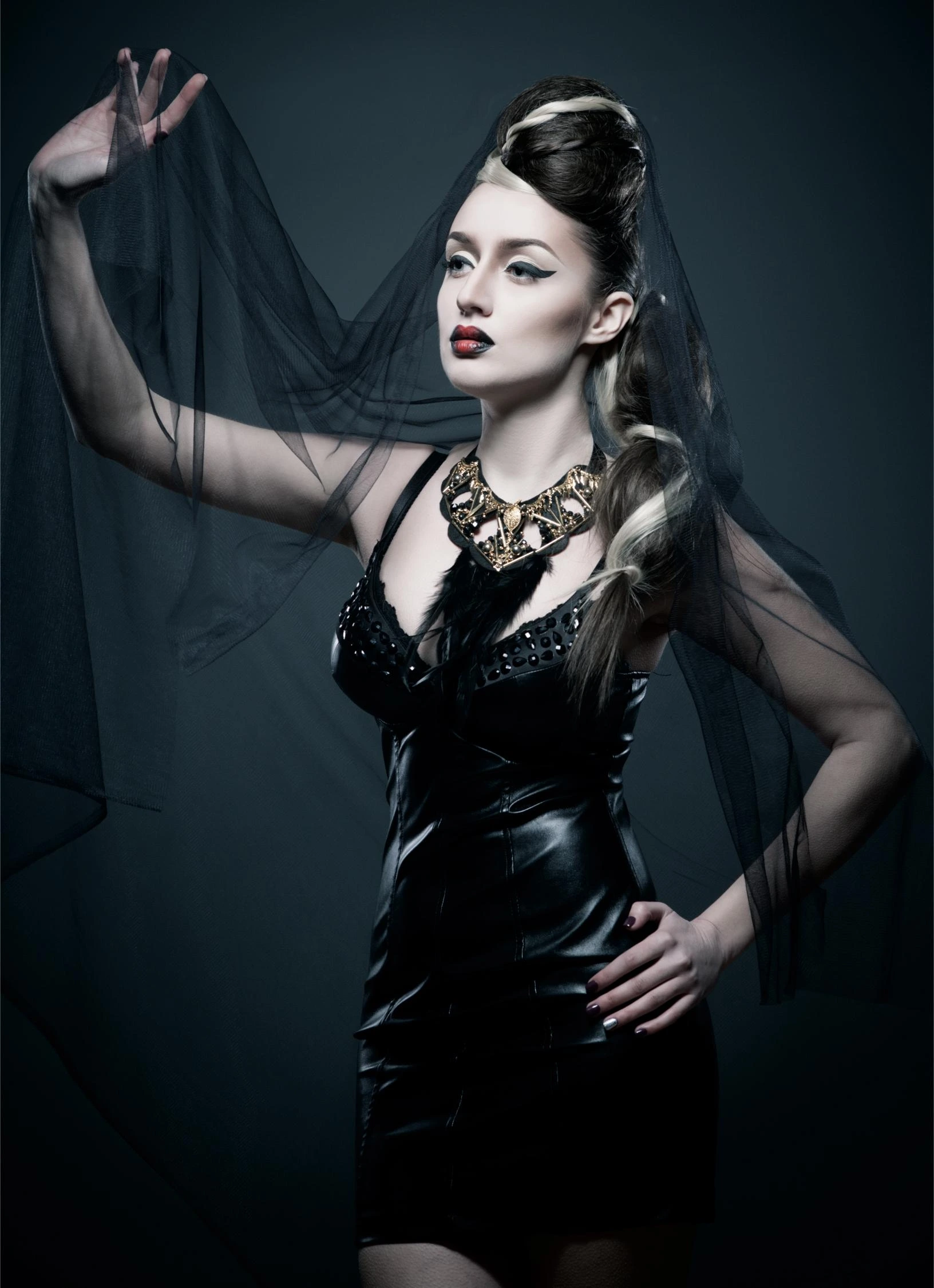 Woman in a gothic veil and a black dress with a statement necklace, posing with an intense expression and red lipstick.
