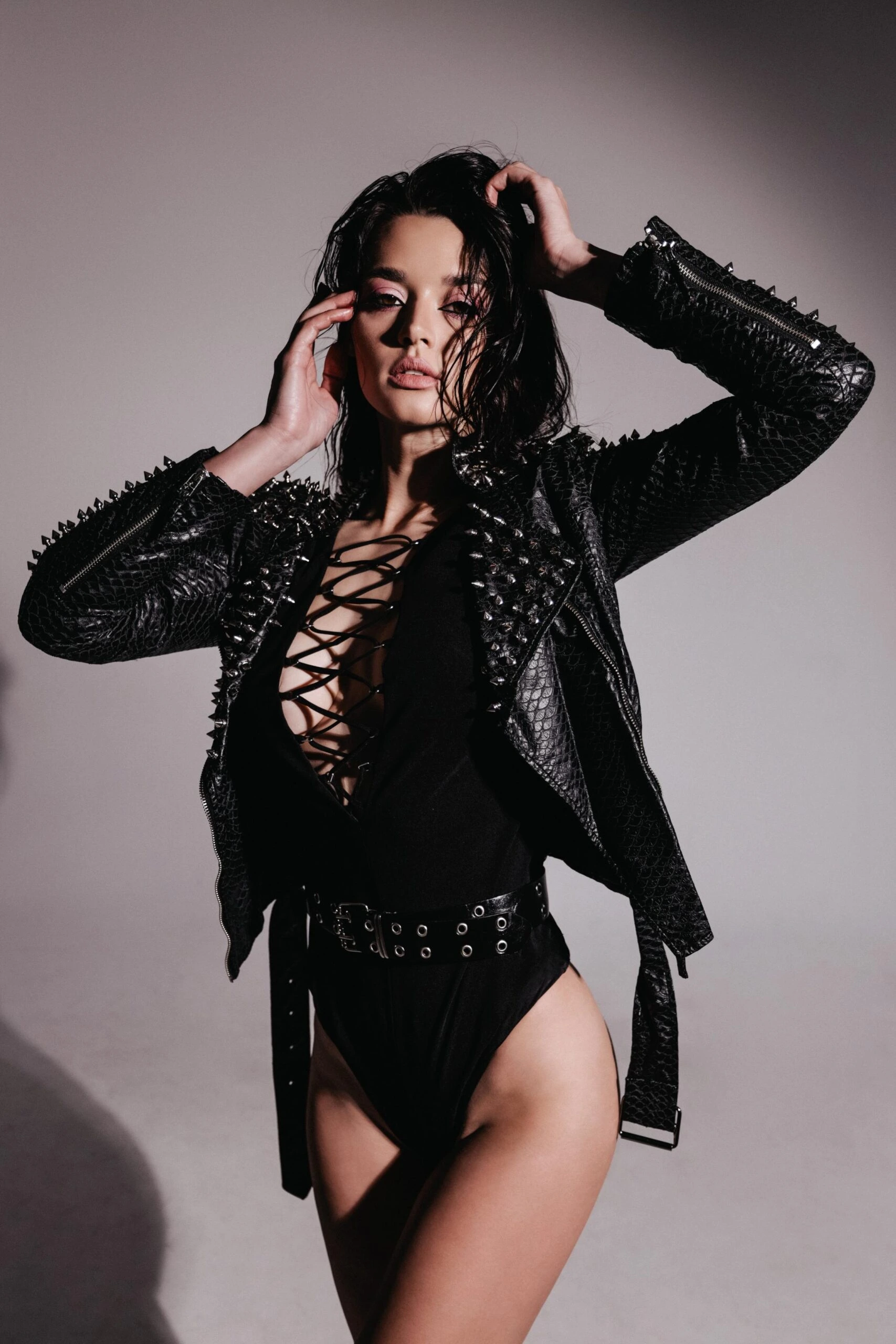 Woman in a studded black leather jacket and a laced bodysuit posing confidently, showcasing edgy goth fashion.