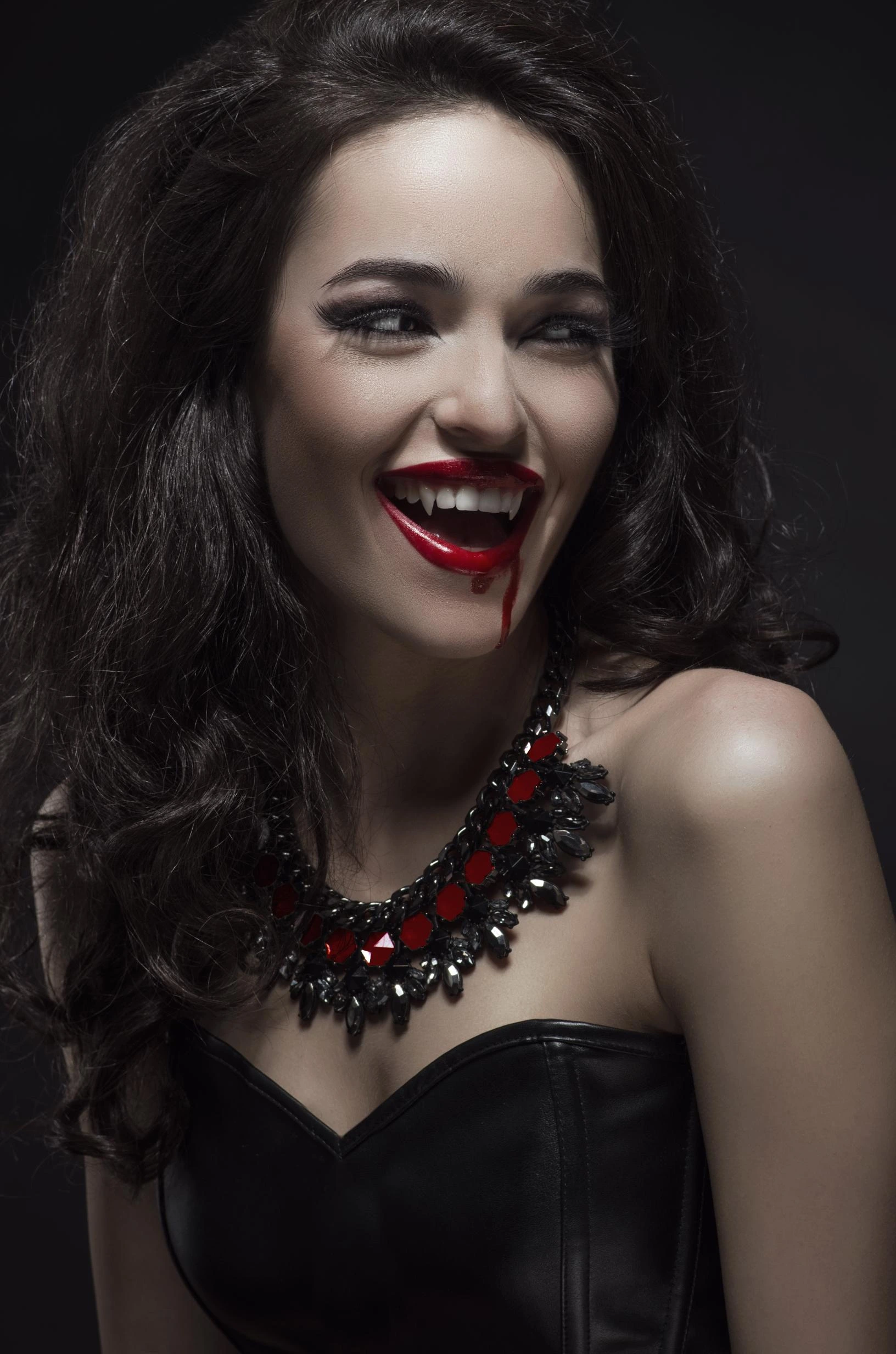 Woman smiling with dark red lipstick and fangs, wearing a black corset and a gothic necklace with red stones.