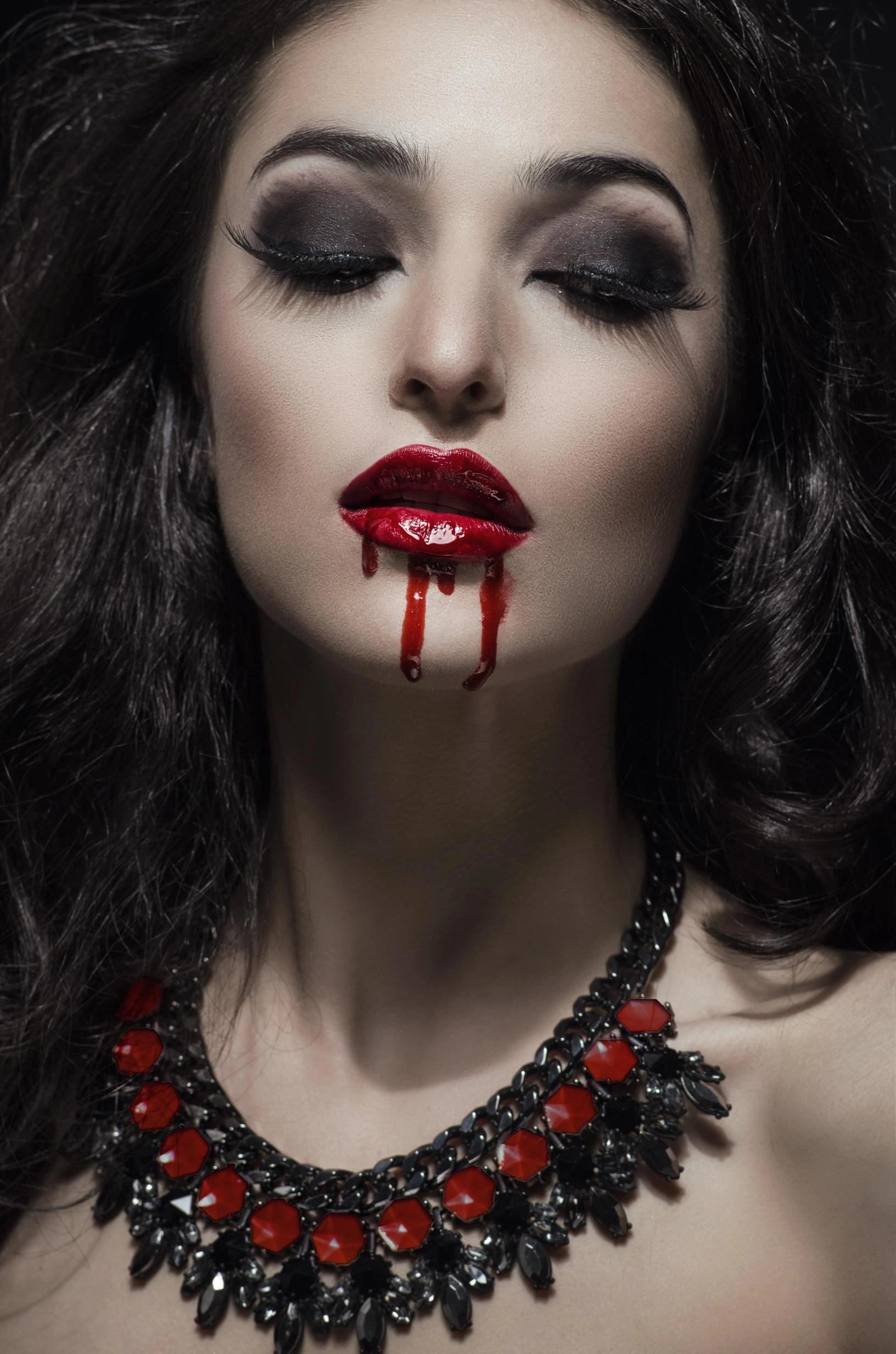 Close-up of a woman with dark makeup and red lips, with blood-like droplets dripping down her mouth.