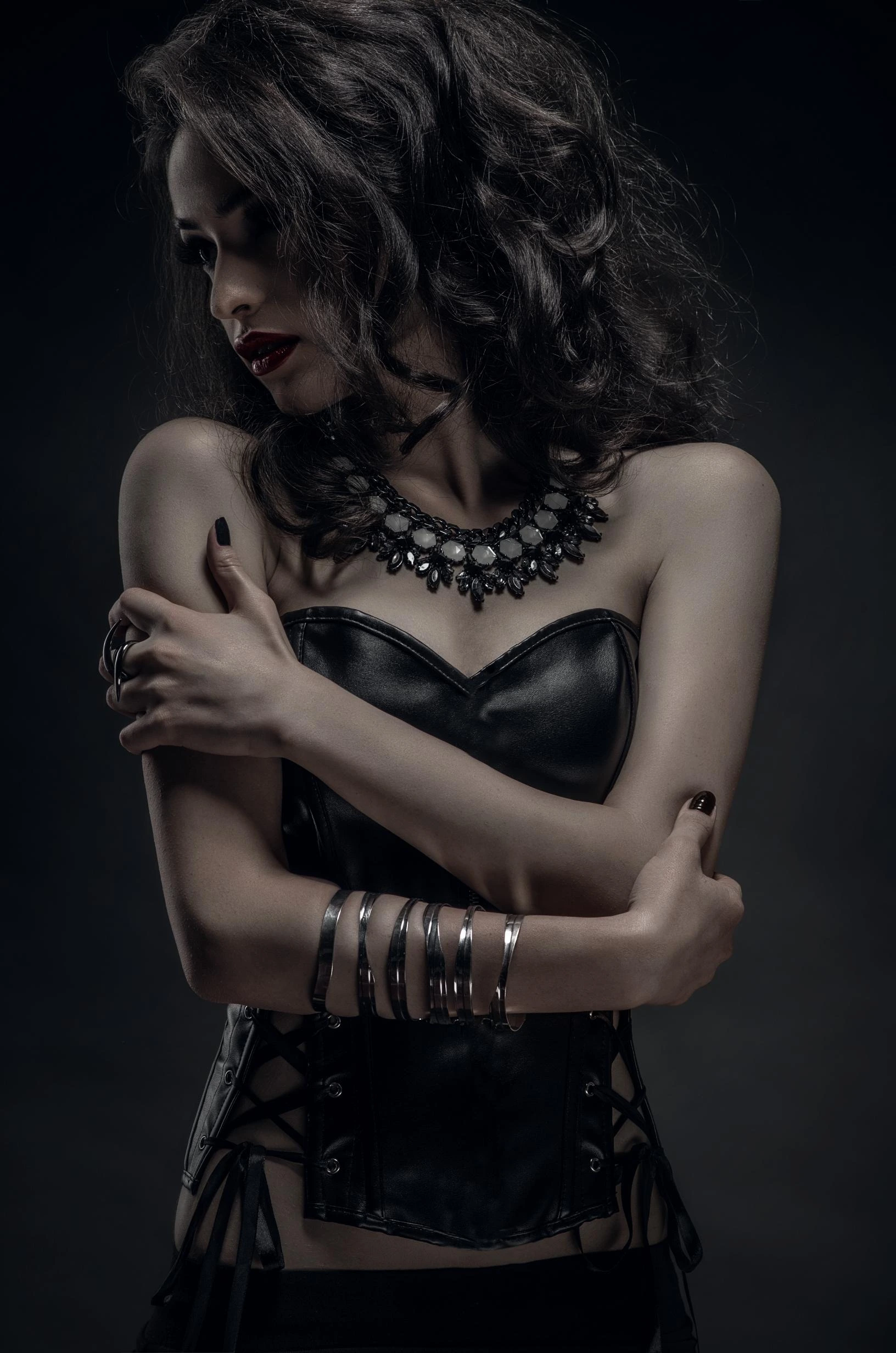 Woman in a corset with arms crossed, wearing dark makeup and a black necklace, gazing downward in a dramatic pose.