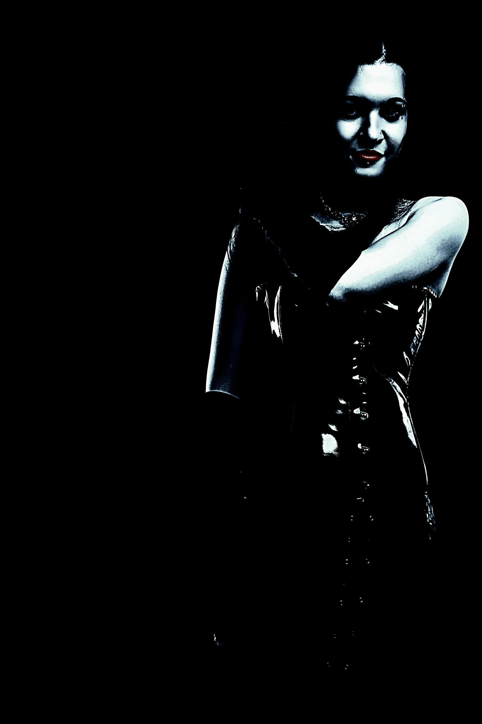 Mysterious woman in a black latex corset dress, partially illuminated in dramatic lighting against a pitch-black background.