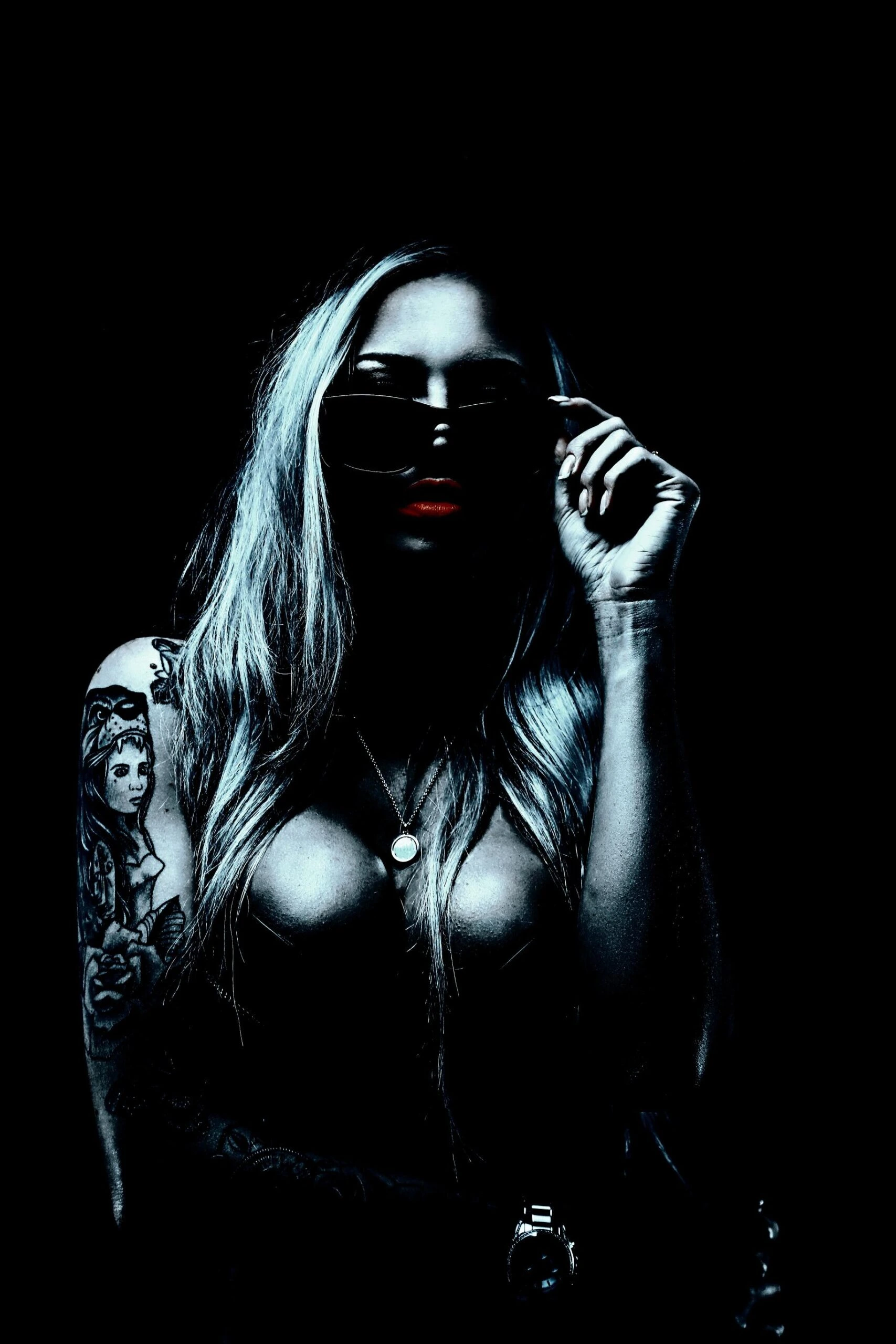 Tattooed woman with sunglasses, red lipstick, and necklace in dramatic lighting, embodying edgy alternative style.