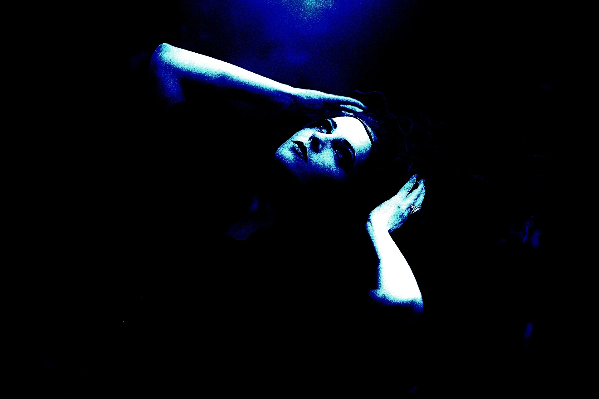 Woman lying back with hands in hair, looking upward in dramatic blue light with a dark, mysterious expression.