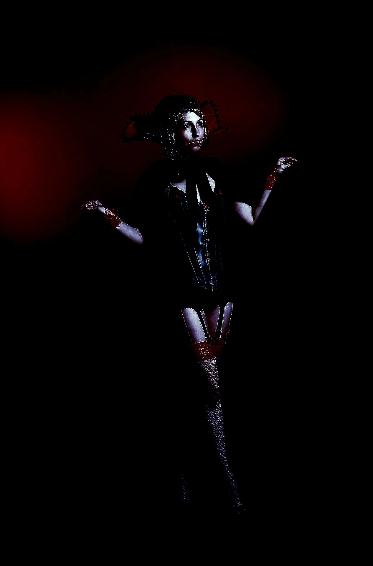Woman dressed in gothic vampire attire with a red and black theme, posed with arms raised against a dark backdrop.