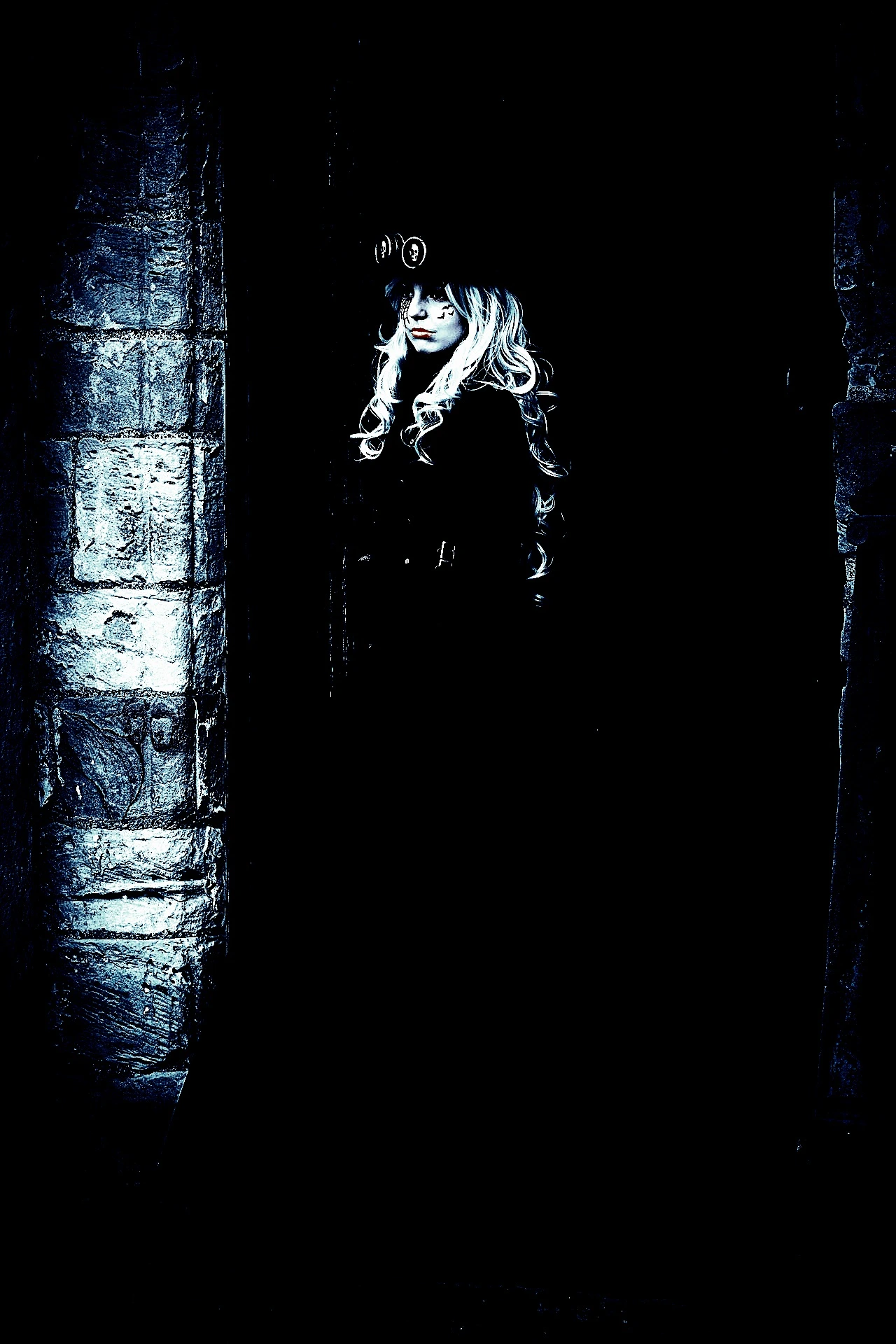 Blonde woman in steampunk-inspired gothic attire standing against a stone wall, blending darkness and vintage style.