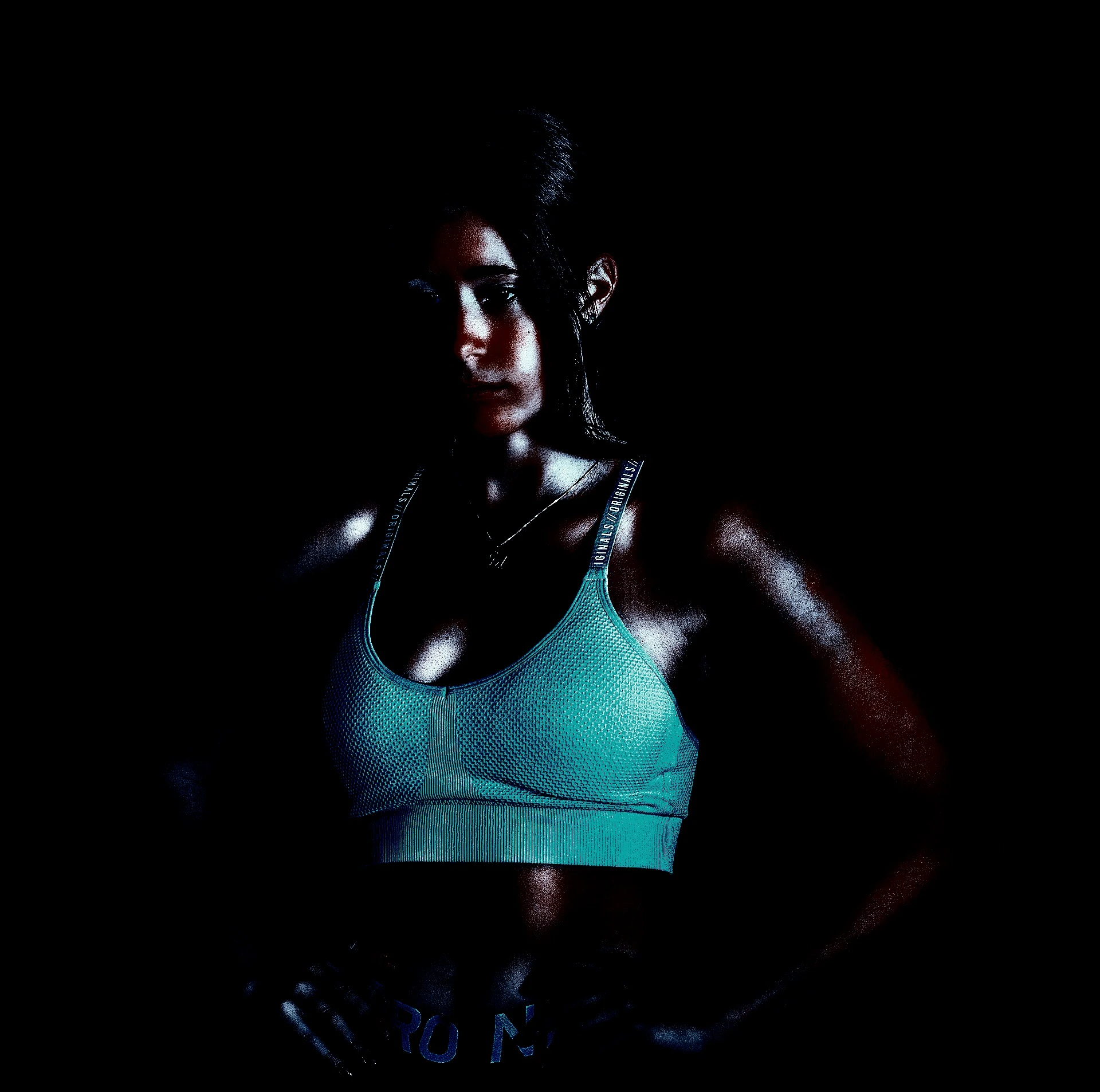 Woman in a teal sports bra standing confidently in low light, with muscles glistening under dramatic lighting.
