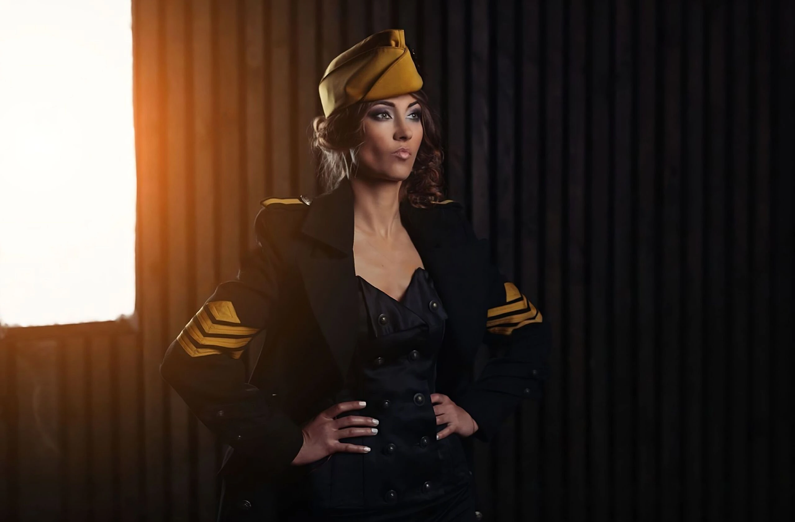 A commanding woman in a dark military-style coat with gold chevron patches and a matching hat, standing with hands on her hips in a dimly lit room.