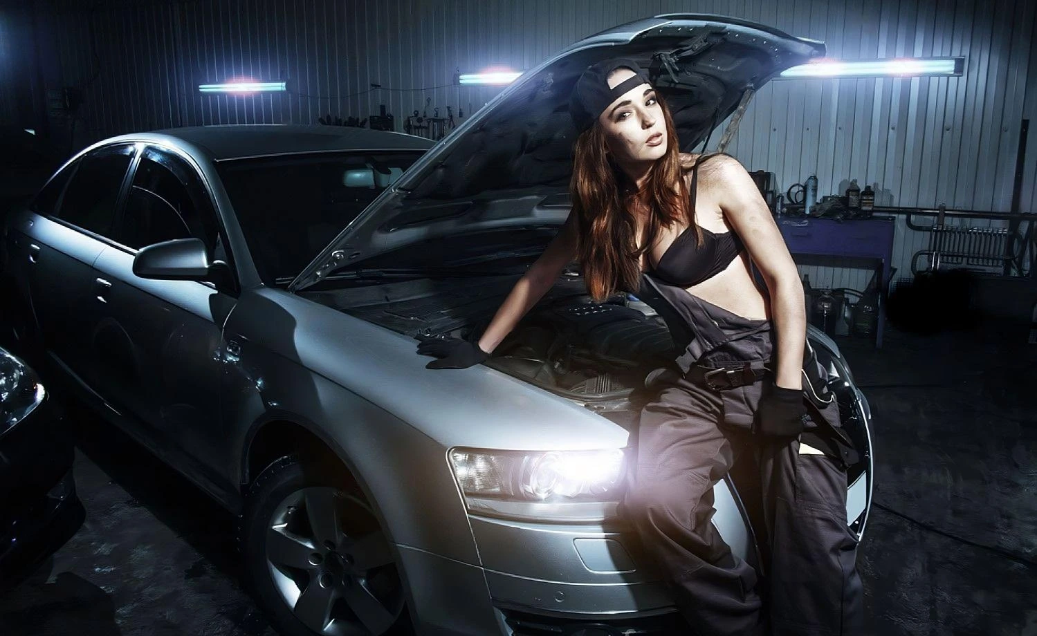 A woman in a mechanic’s jumpsuit and black bra leans against a car with its hood open in a dimly lit garage.