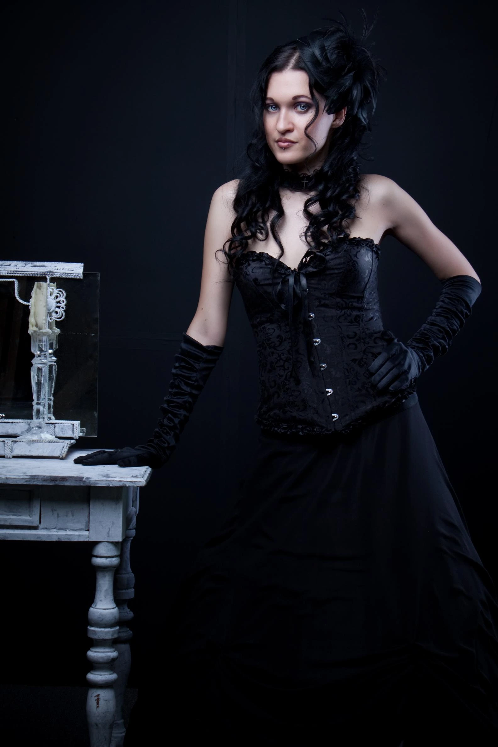 Elegant woman in a black corset dress with intricate lace details, standing beside a vintage table adorned with a candleholder.