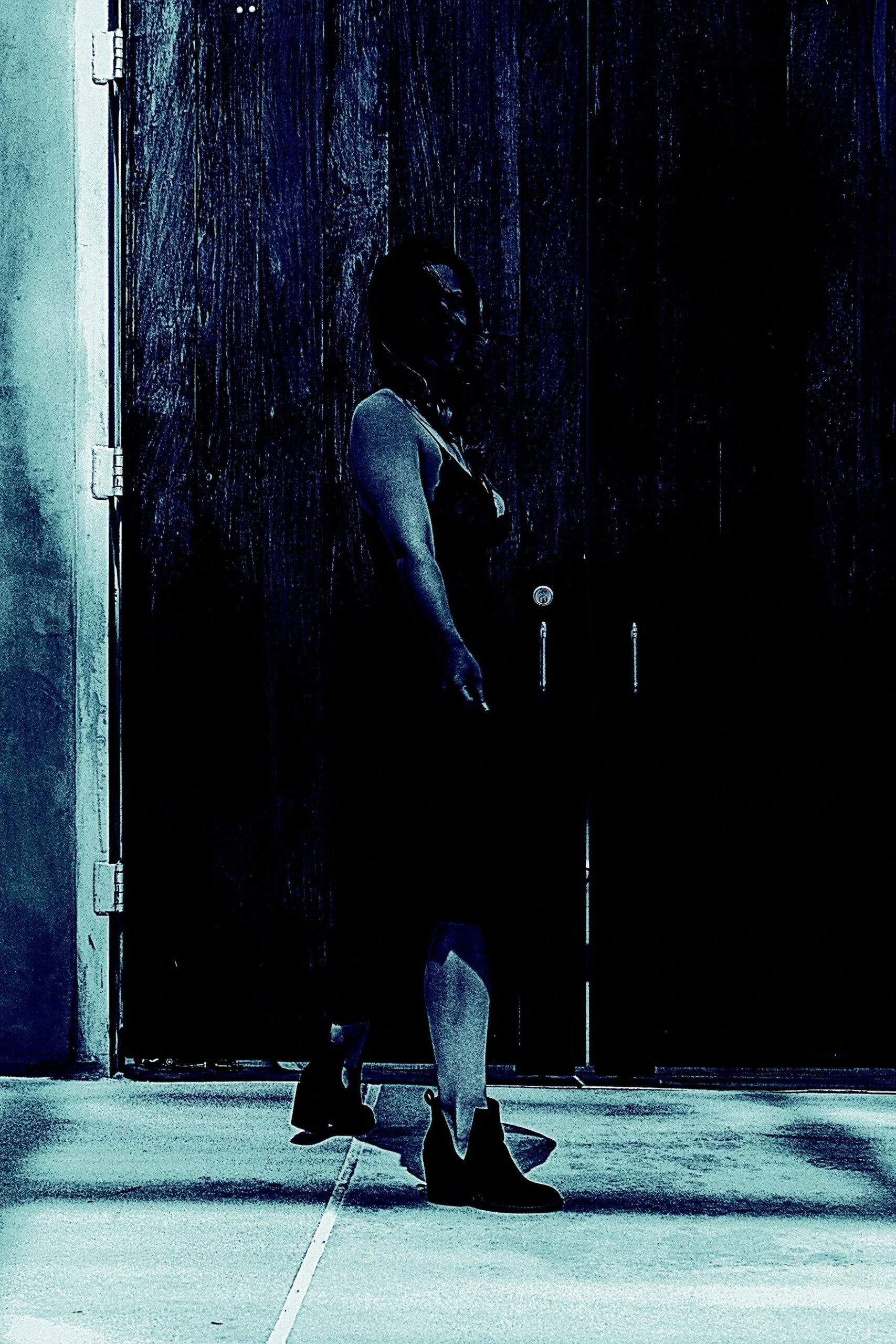 Silhouetted woman standing against a large, dark wooden door, partially lit with an eerie blue hue.
