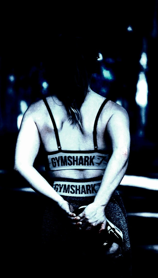 Back view of a woman in Gymshark workout gear, holding a resistance band, standing in a dimly lit setting with a dark, moody vibe.