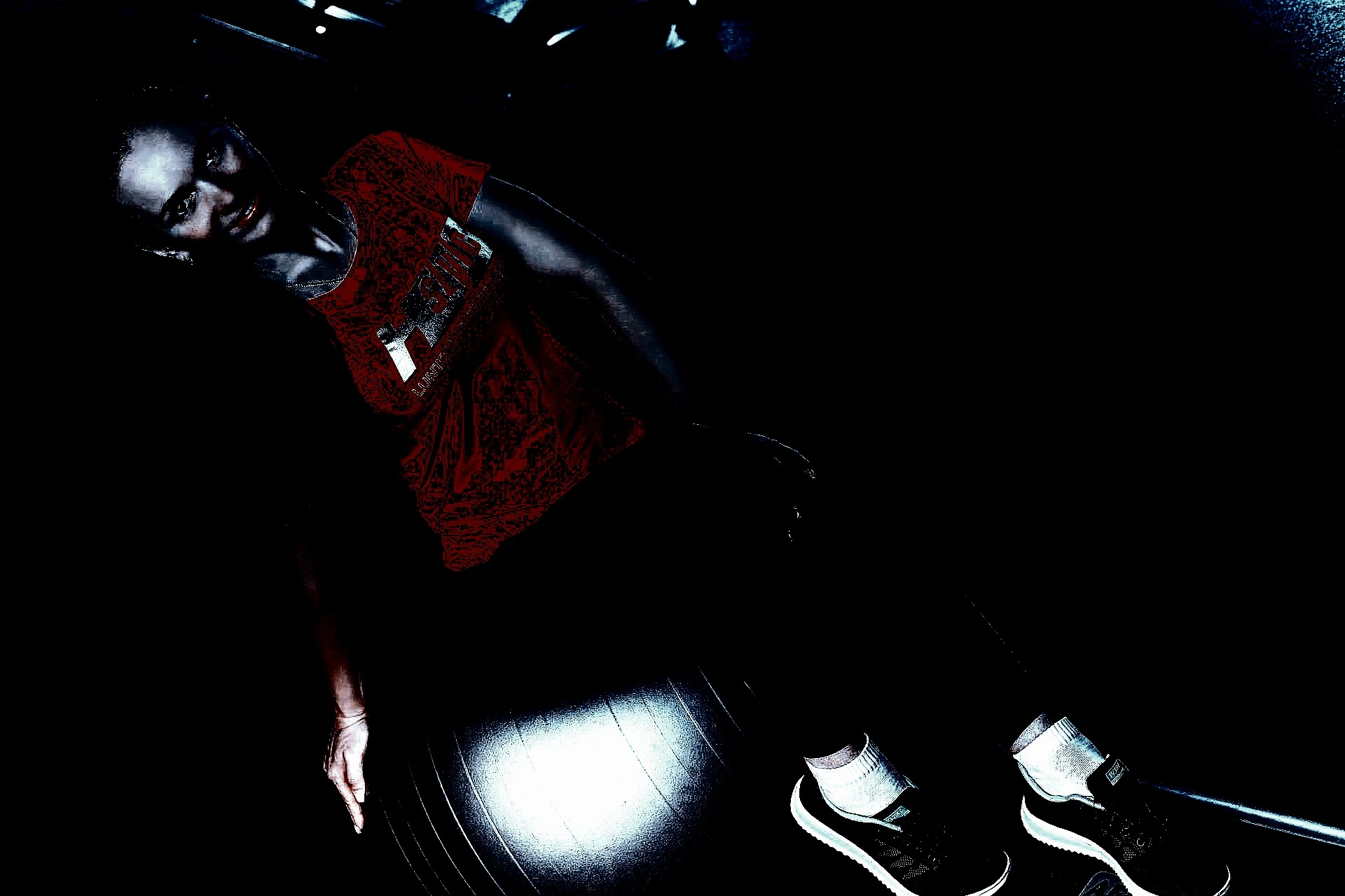 Focused woman seated on an exercise ball in a dark gym, wearing a red shirt with metallic lettering, illuminated by subtle lighting.