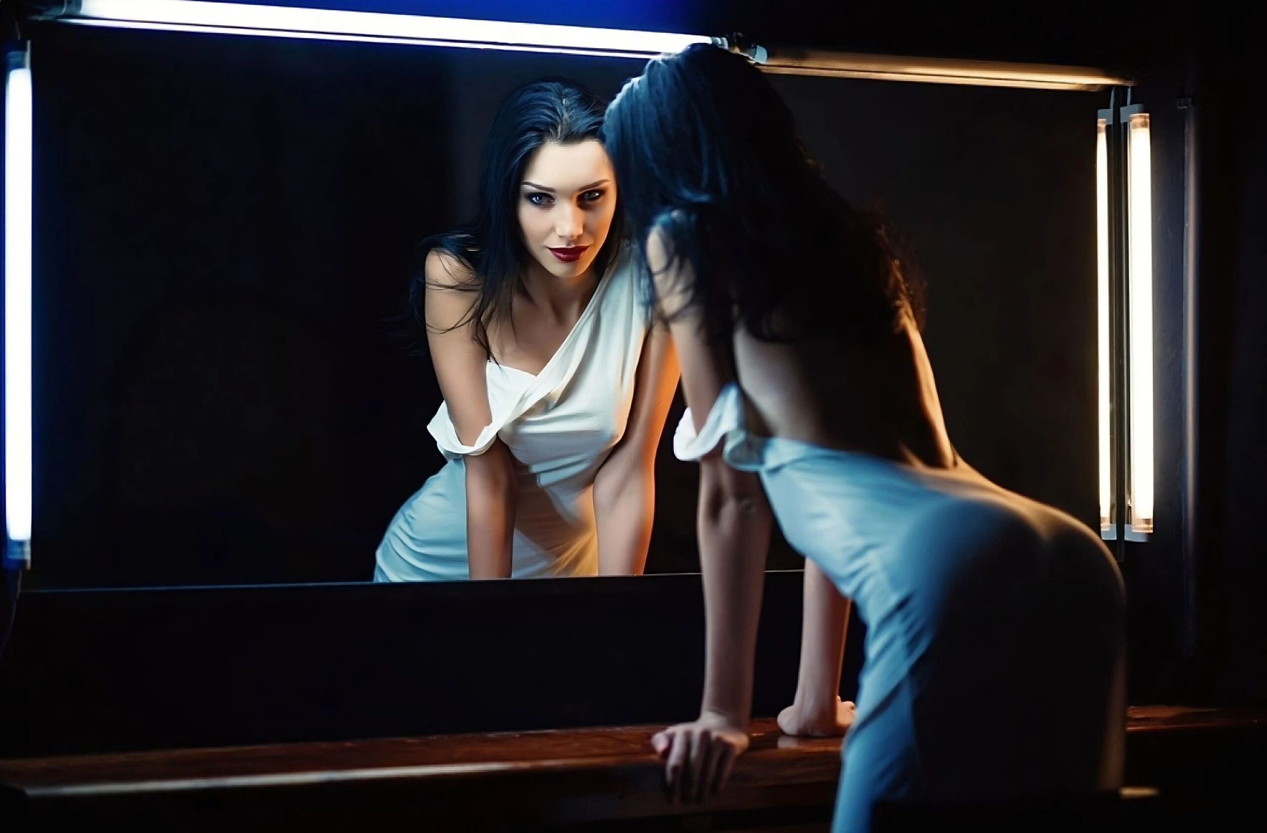 A glamorous woman in a white dress leaning against a mirror, illuminated by soft lighting that creates a seductive vibe.