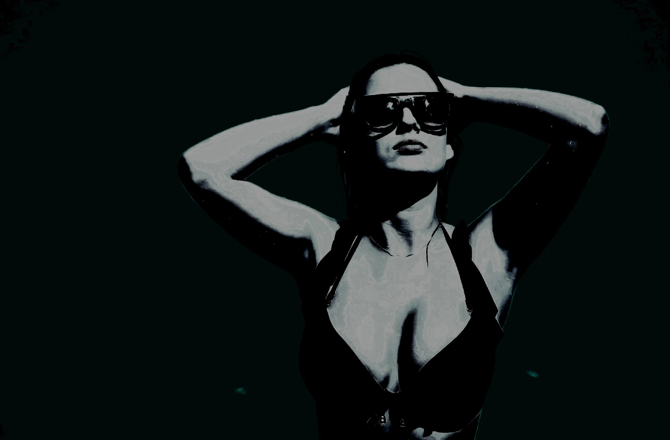 A woman in a black bikini and oversized sunglasses, confidently holding her hands behind her head against a shadowy background.