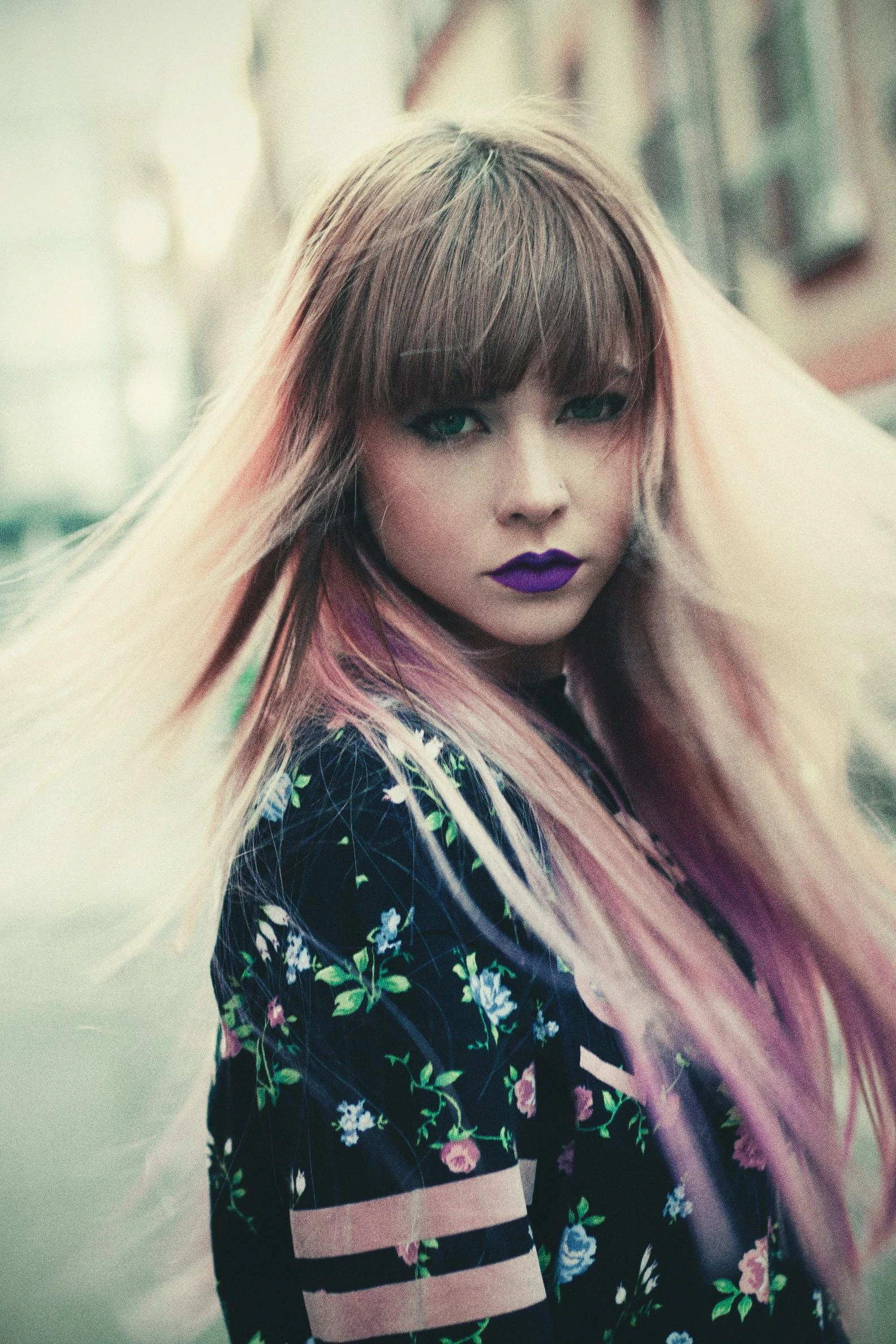 A woman with pastel hair and dark makeup, wearing a floral streetwear outfit, looking into the camera with intense energy.
