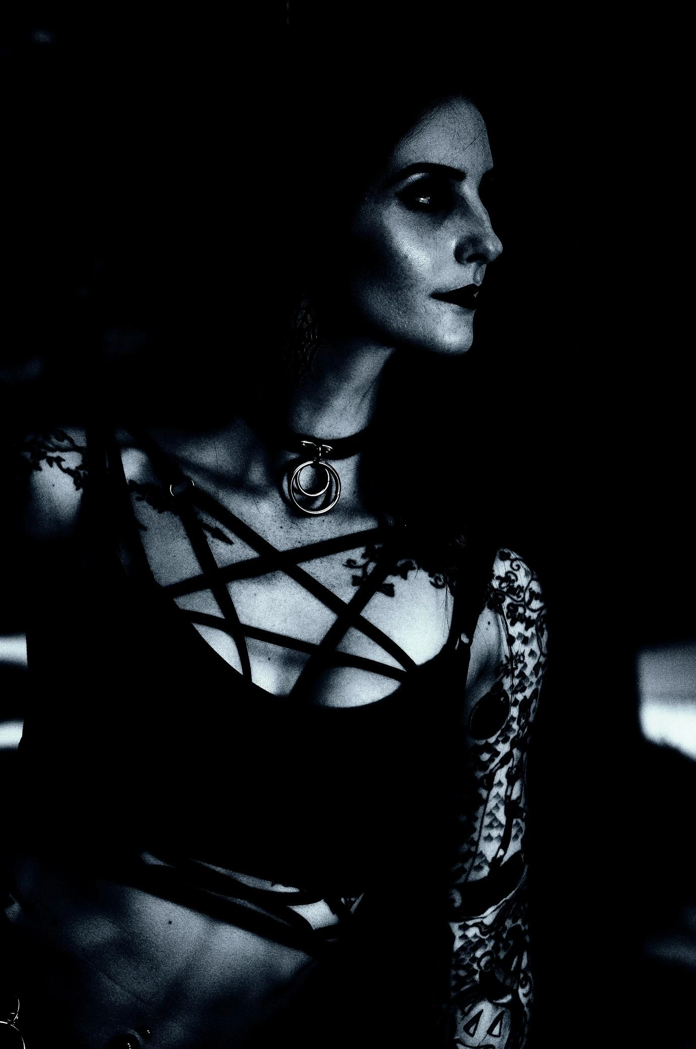 A gothic woman wearing a pentagram harness top and choker, gazing into the distance with dark makeup and tattoos.