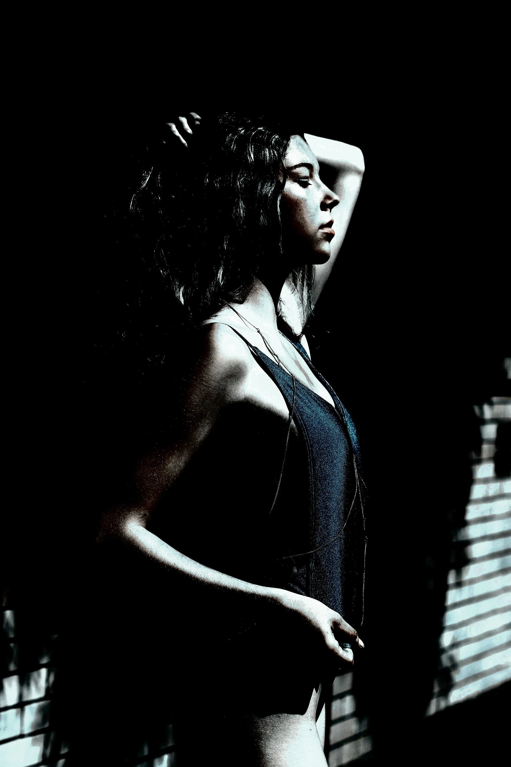 A woman in a moody, high-contrast setting, posing with one arm raised, wearing a dark minimalist outfit.
