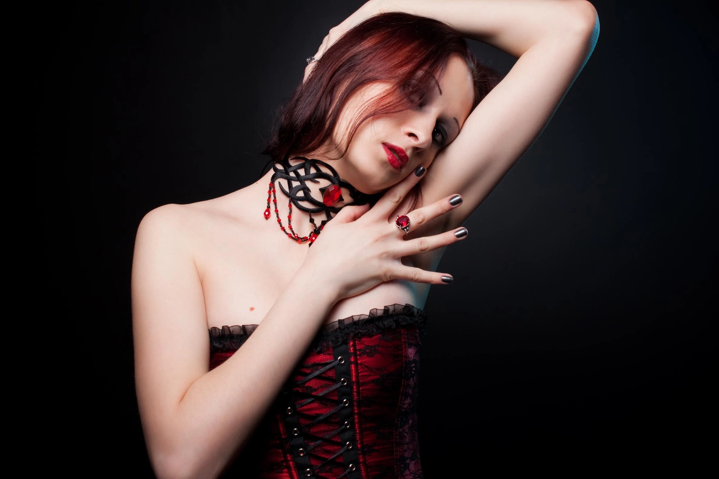 Woman in a red corset with intricate choker, gazing intensely against a dark backdrop, epitomizing gothic fashion.