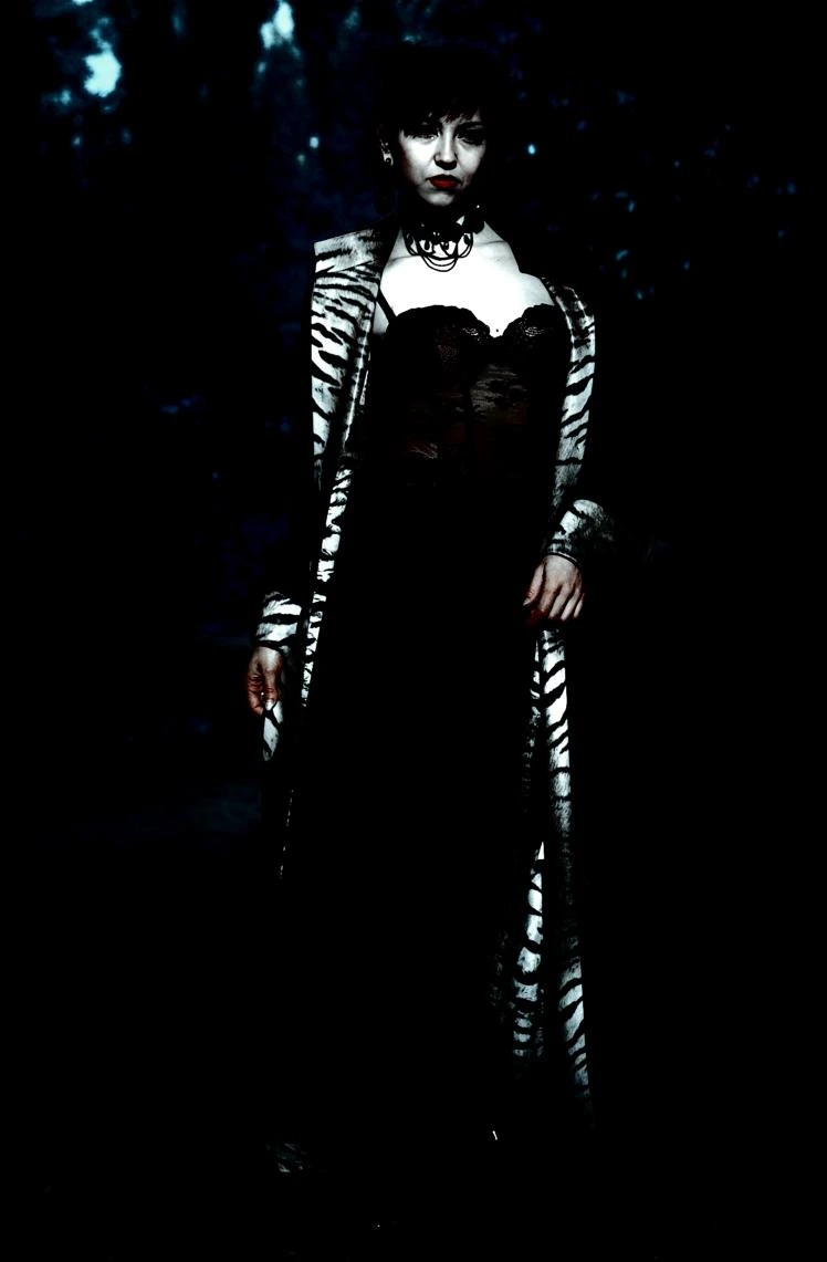 Woman in a zebra-print coat over a sheer lace bodice, standing in a dark, eerie forest, blending elegance with darkness.