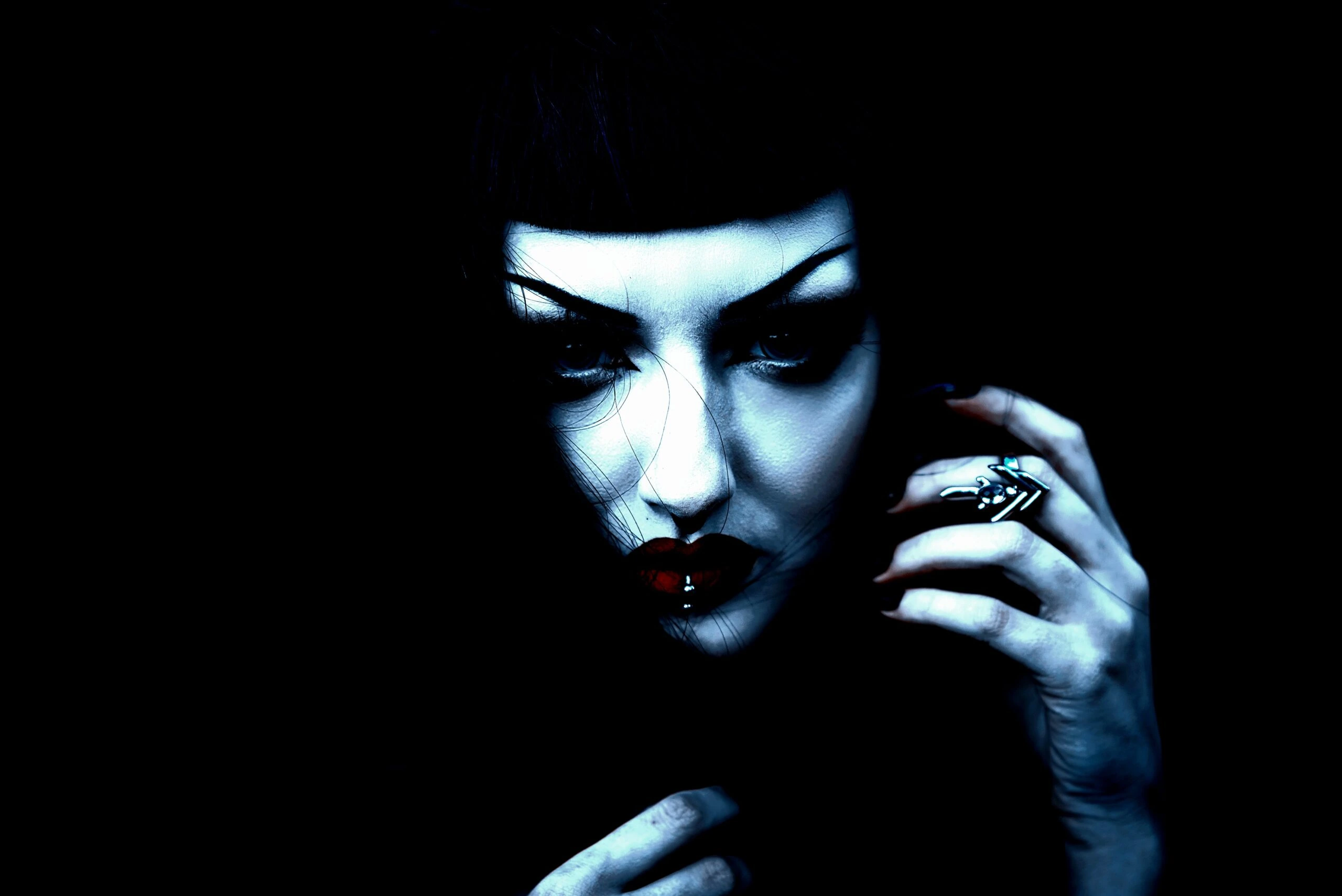 A gothic woman with dramatic makeup, dark arched eyebrows, red lips, and a lip piercing, gazing intensely in a moody, high-contrast portrait.