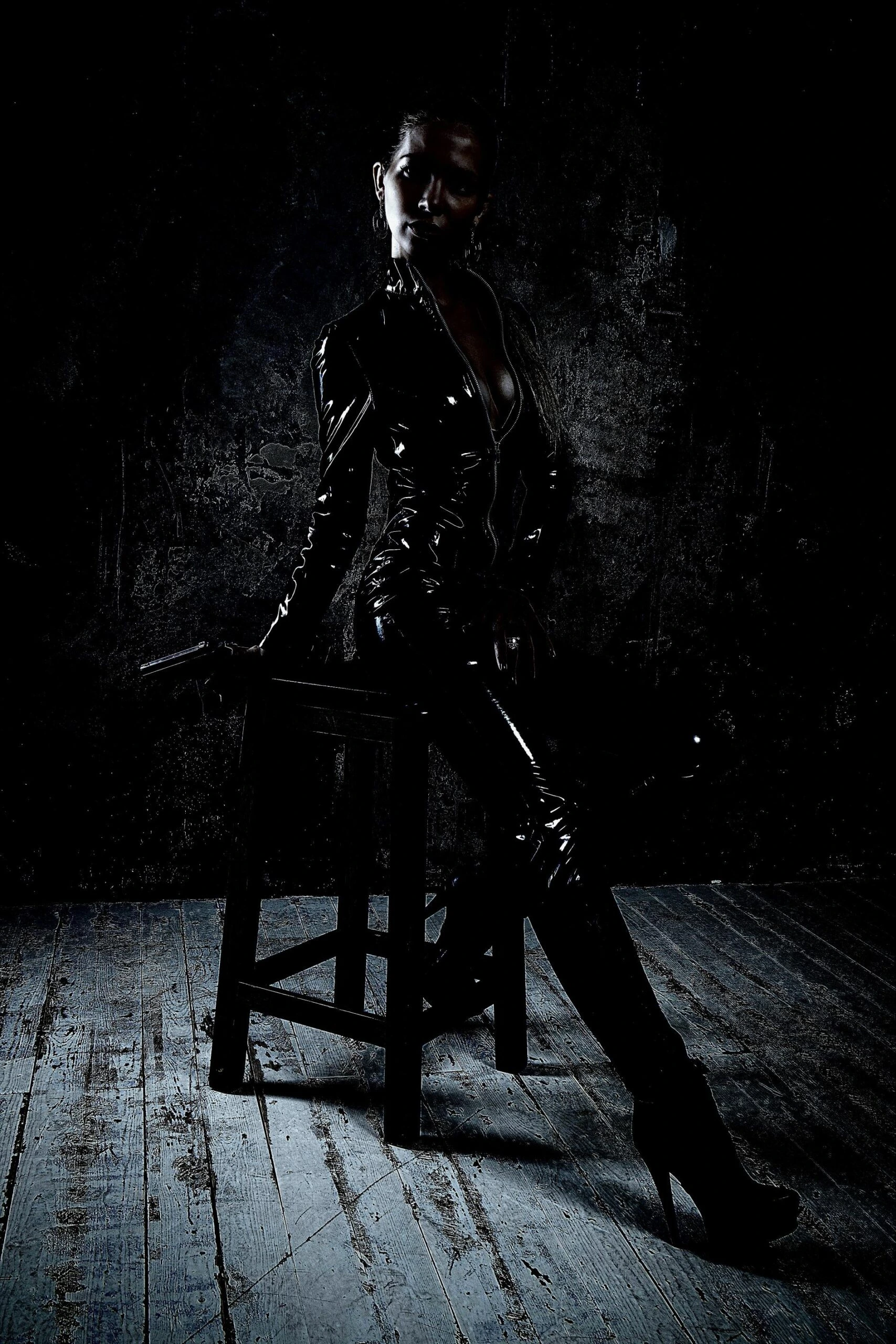 Confident woman in a black latex catsuit and high heels, sitting on a stool in a grungy, dimly lit room.
