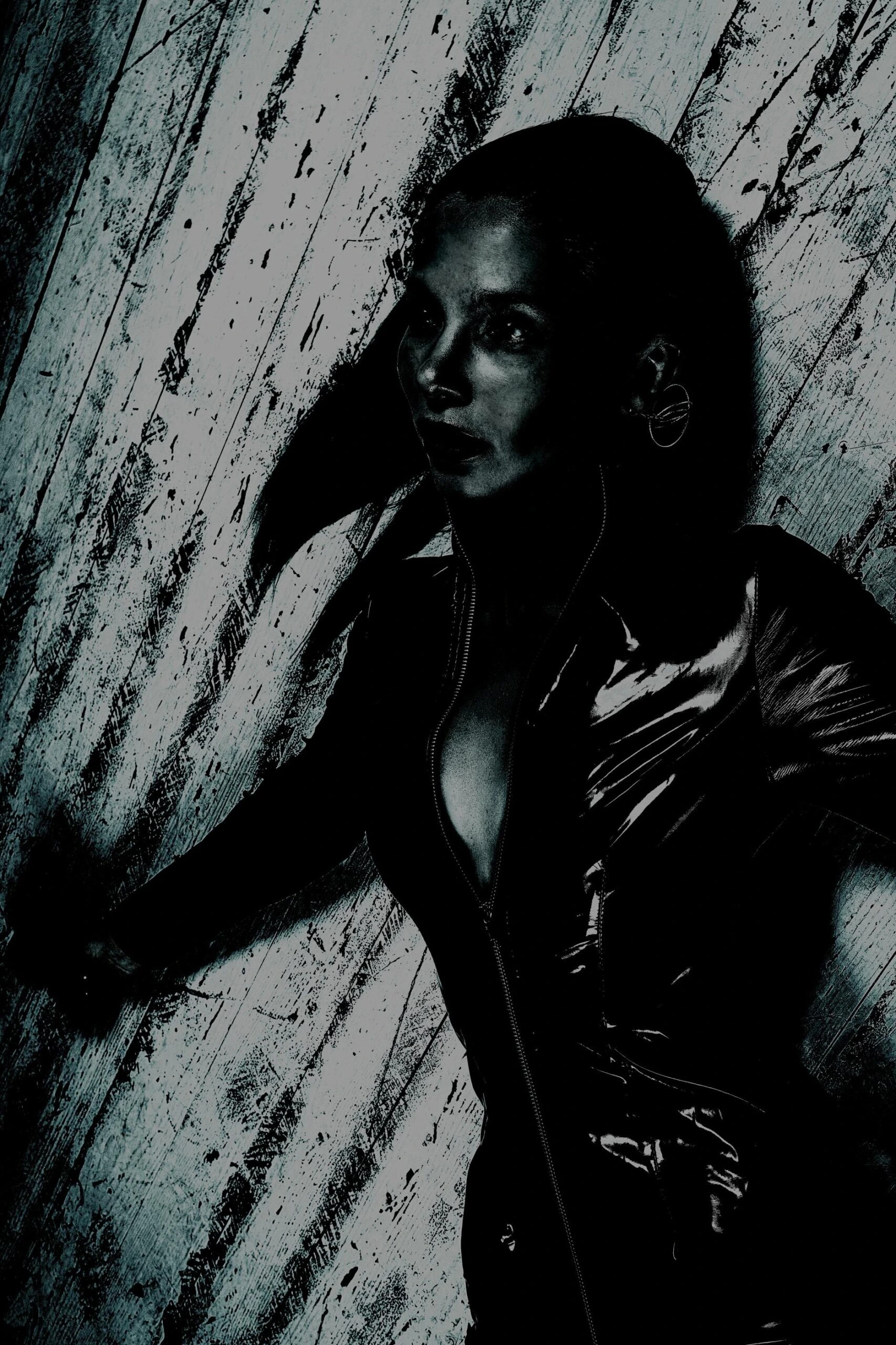 Intense woman in a partially unzipped black latex suit, leaning against a distressed wooden wall.