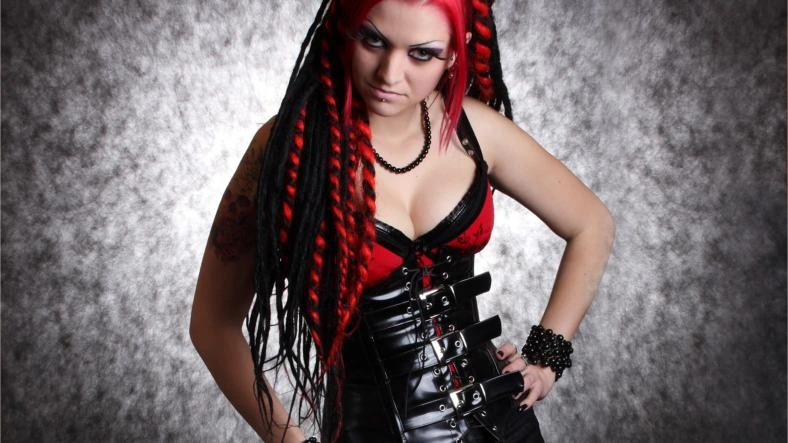 Gothic woman in a tight black leather corset with metal buckles, red lace bra, and black dreadlocks, striking a dominant pose.