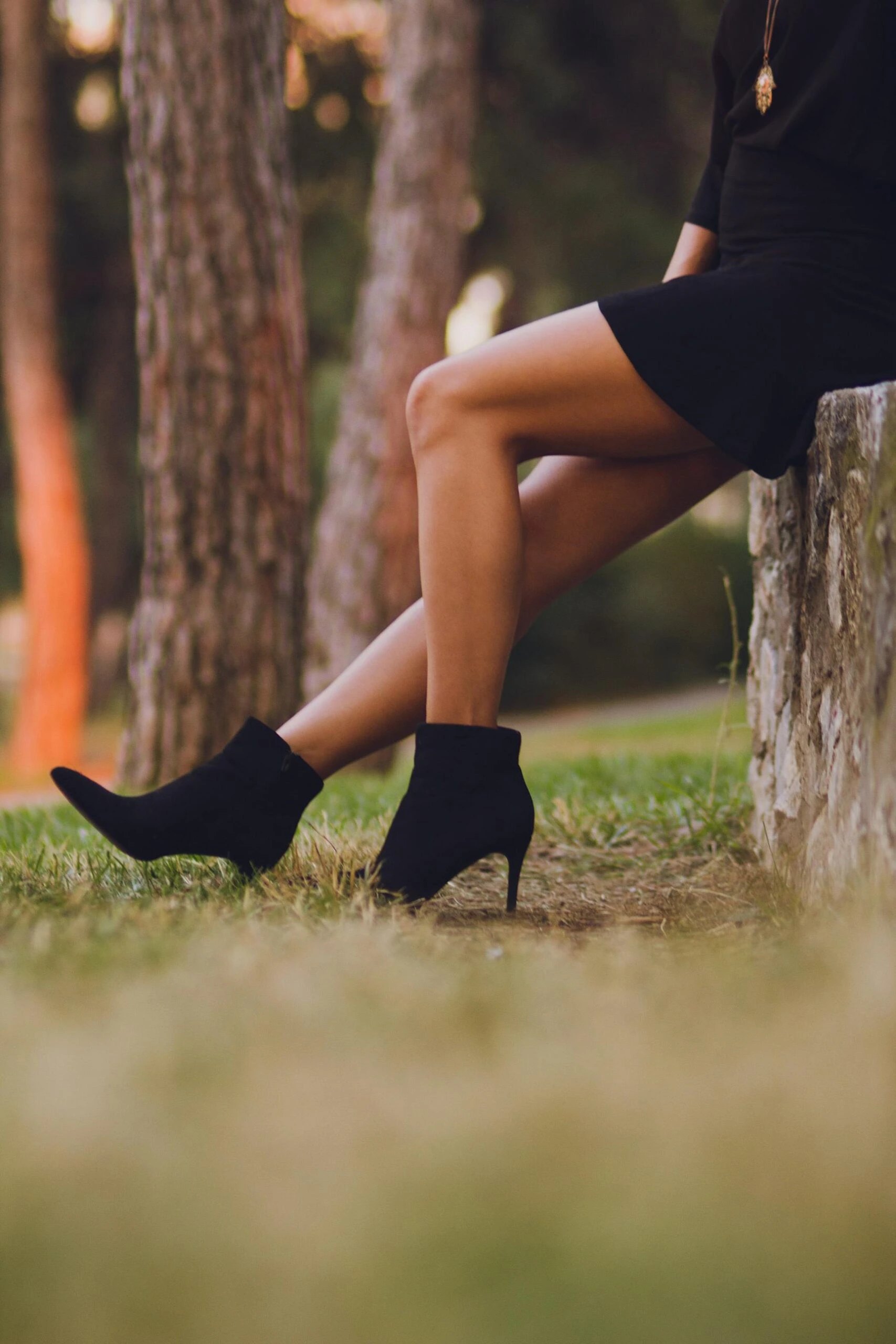 Woman in short black dress wearing pointed black ankle boots sitting on stone edge