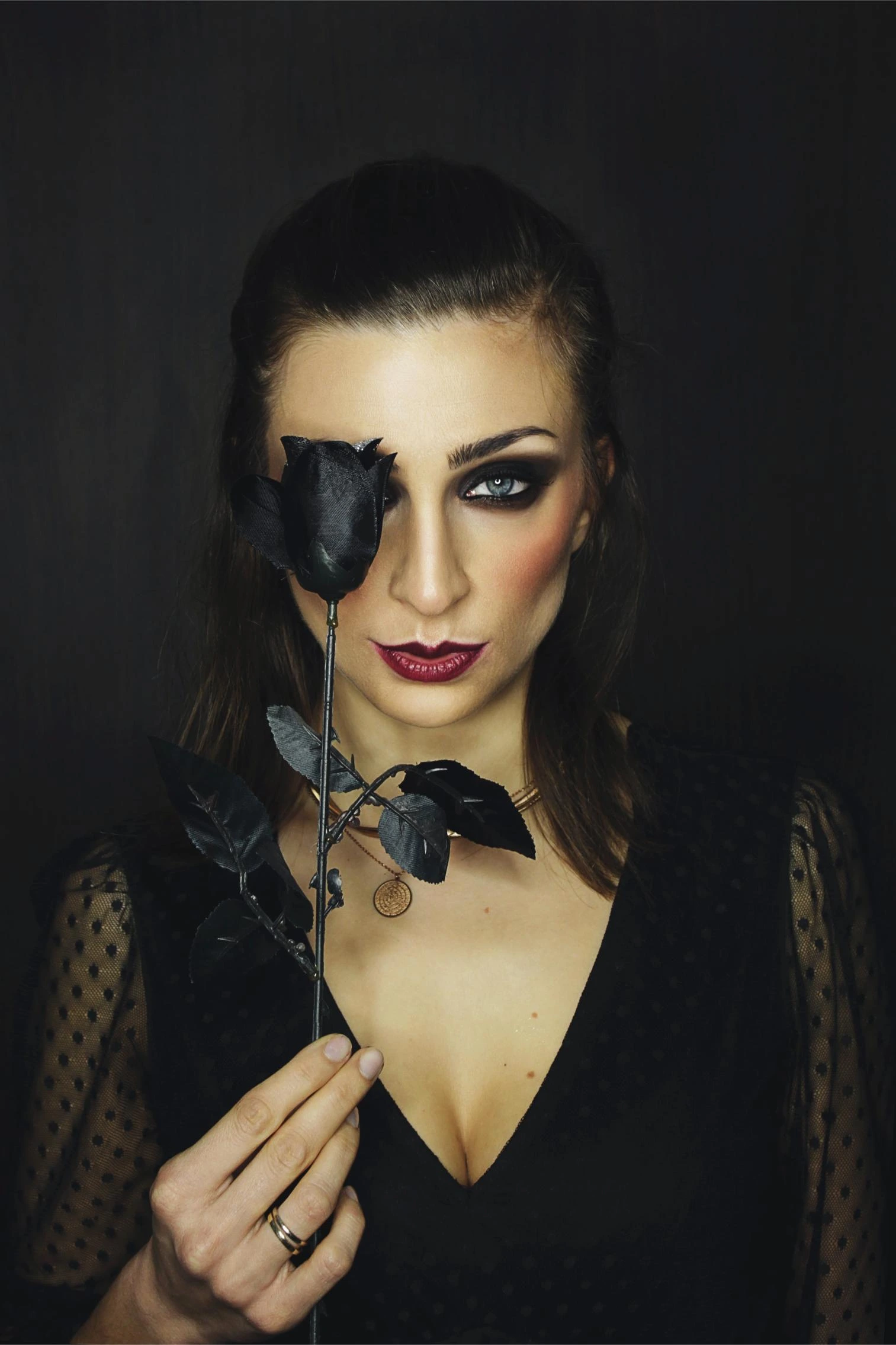 Woman with smoky eyeshadow holding a black rose over one eye, wearing a sheer polka dot black top