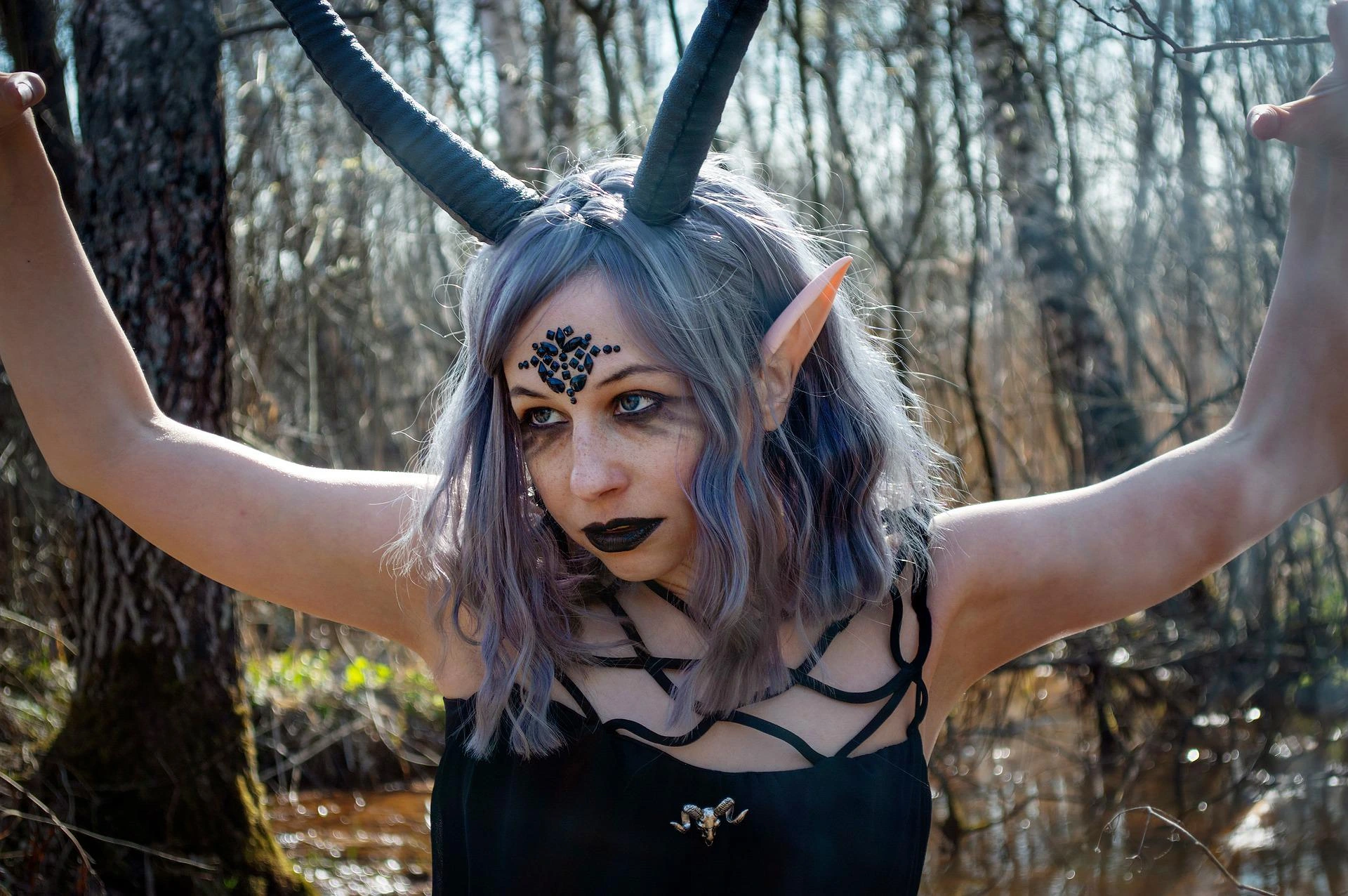 Fantasy woman with long horns, elf ears, and dark makeup in a black strappy dress in forest