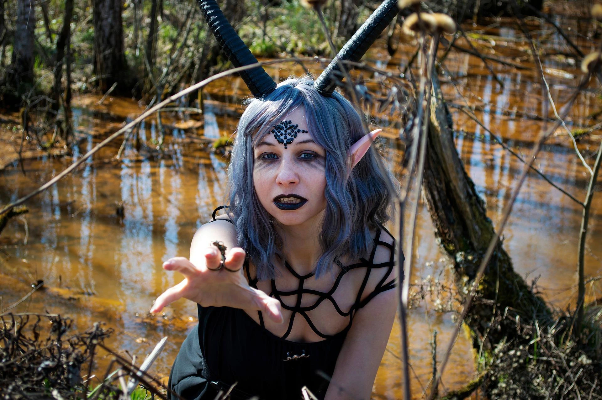 Gothic elf-like woman with horns and black lips reaching out in eerie swamp environment