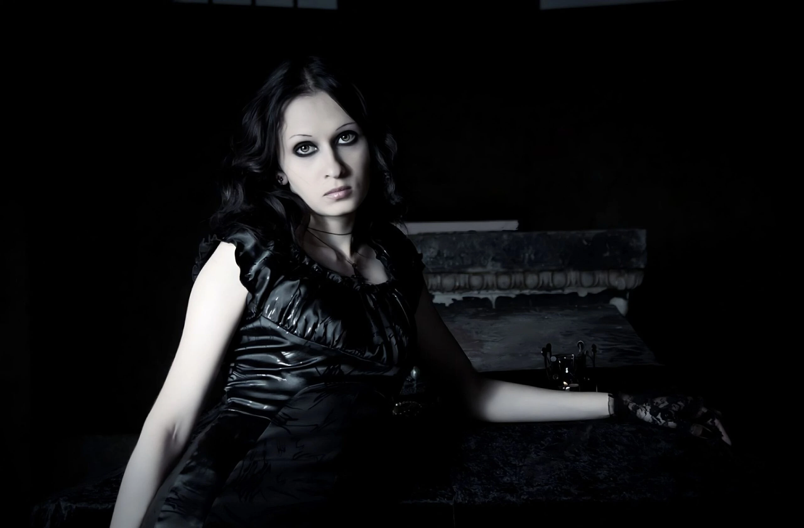 Gothic woman in black latex dress with dark eye makeup in a shadowy room