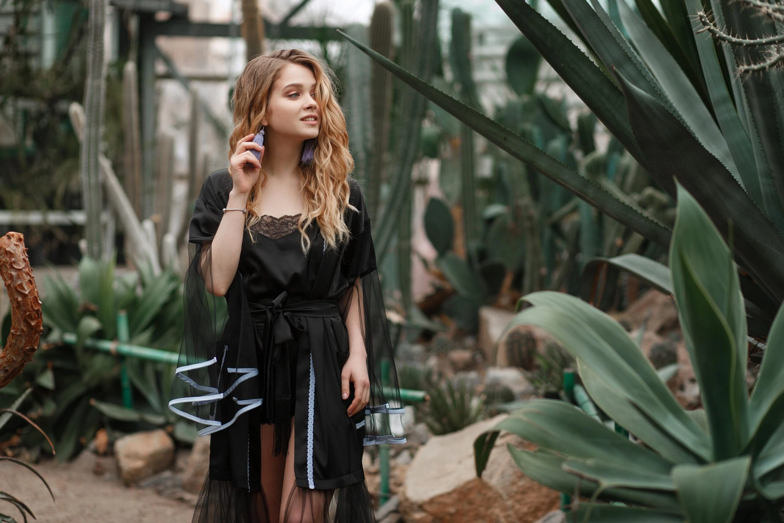 Woman in a black lace-trimmed robe standing in a greenhouse, surrounded by cacti and greenery.