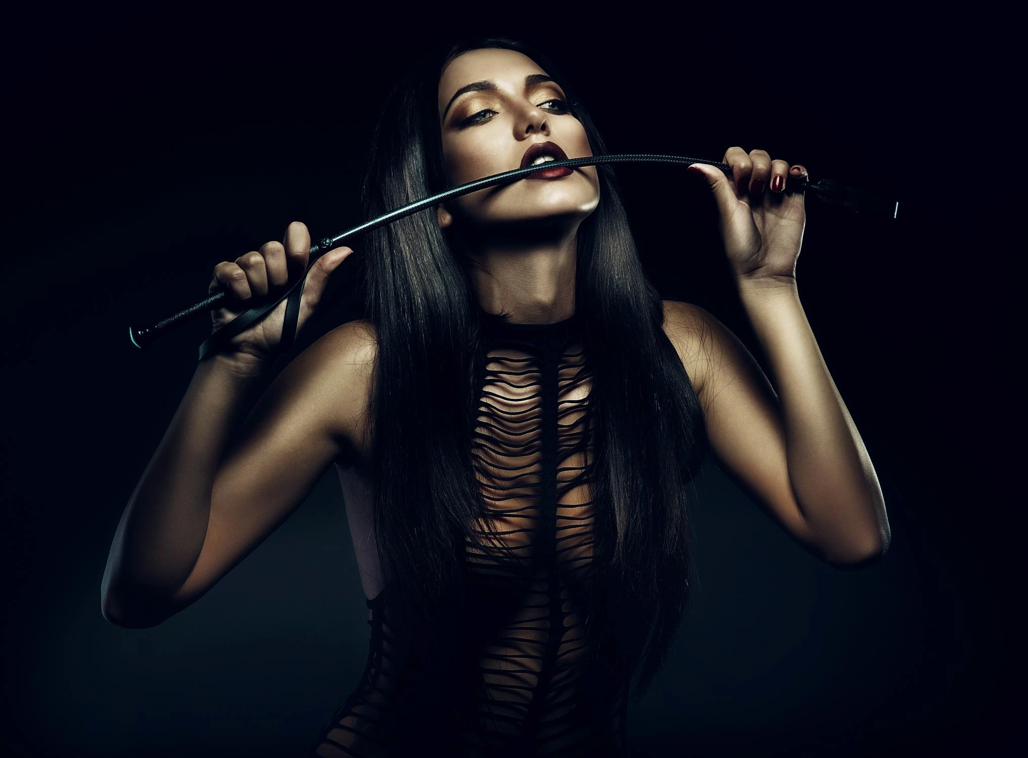Woman with long dark hair holding a riding crop in mouth, wearing black shredded outfit in dramatic lighting